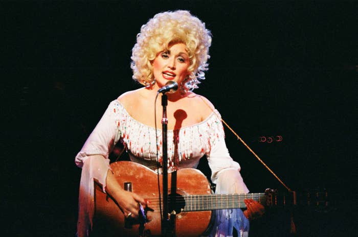 Person performing on stage with a guitar, wearing an off-the-shoulder, embellished dress and a voluminous hairstyle