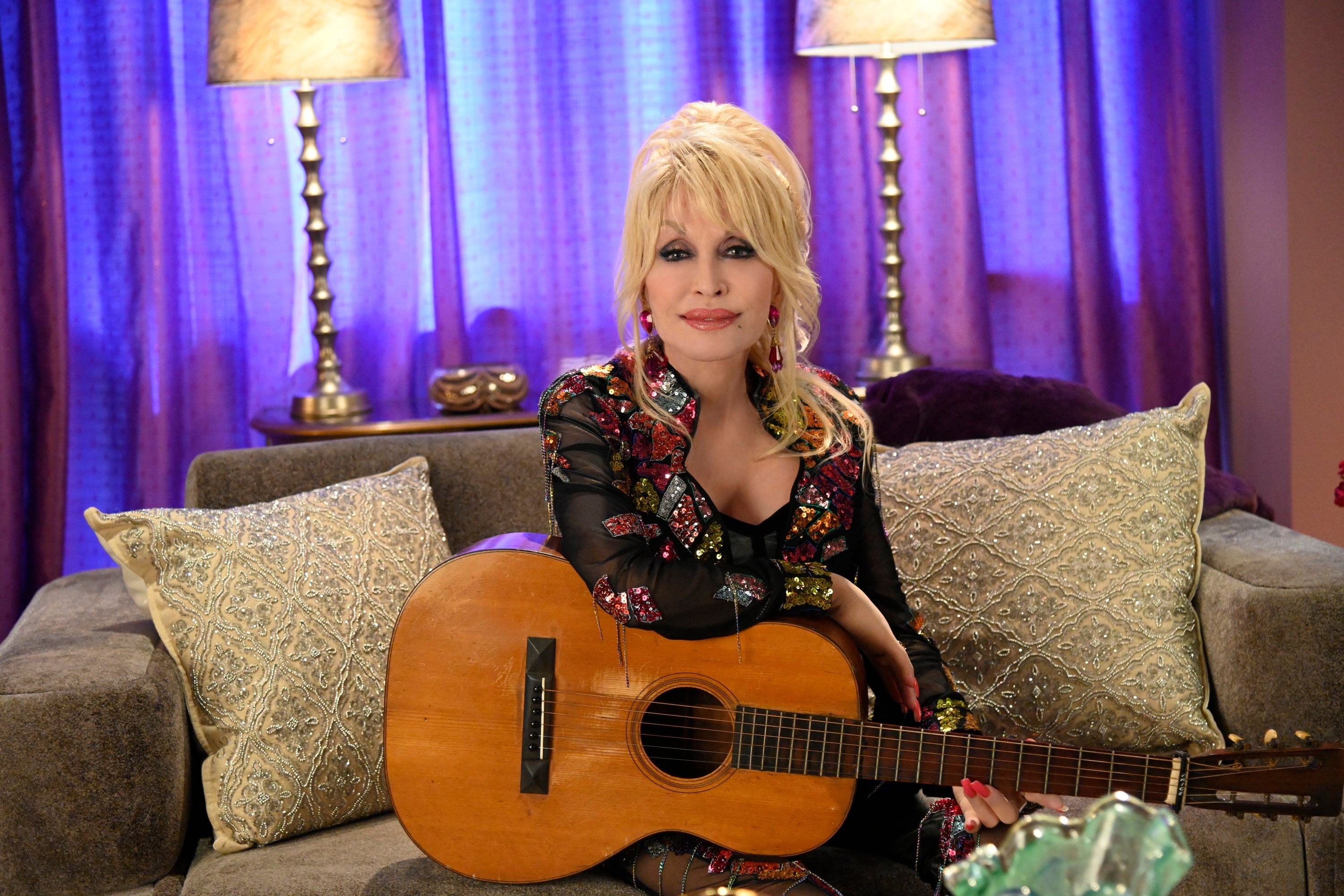 Person sitting on a couch with a guitar, wearing a floral, embellished outfit