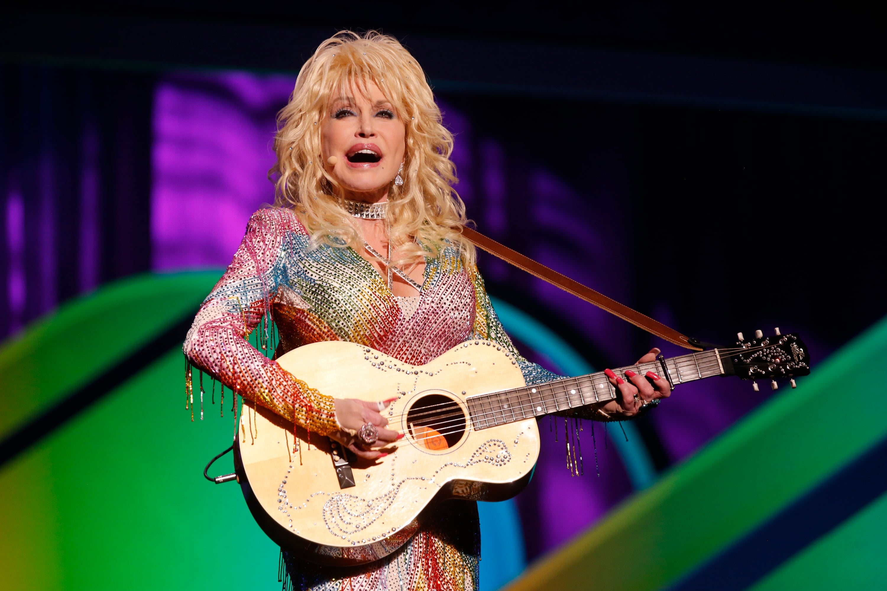 A musician with long, curly hair performs on stage, wearing a sparkling, multicolored outfit, and playing an acoustic guitar
