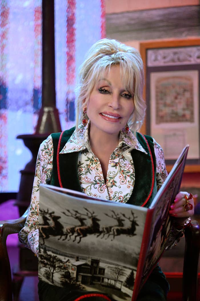 Person with styled hair in an embroidered blouse and velvet vest sits reading a book featuring reindeer illustrations, smiling warmly at the camera