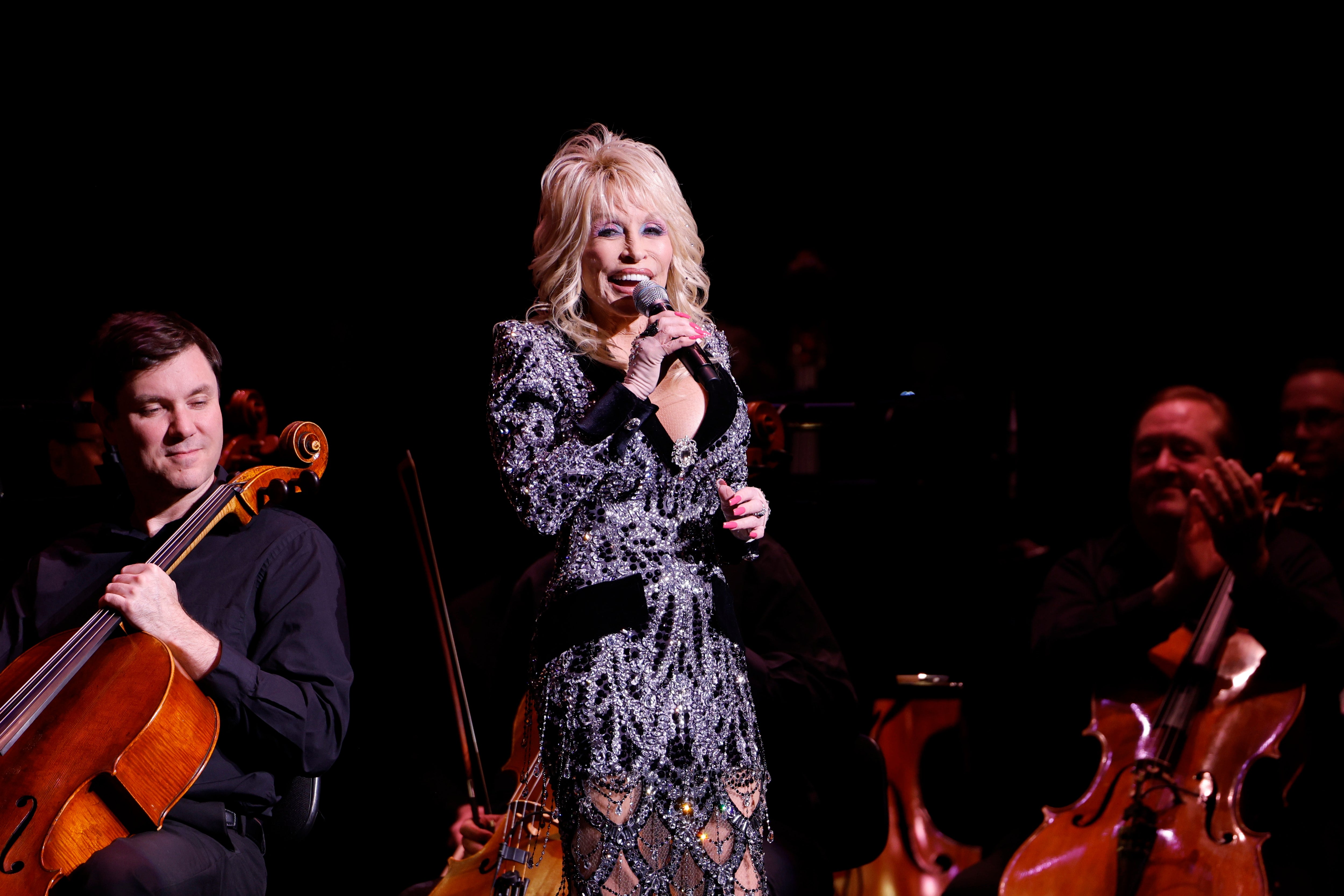 Singer performing on stage in an embellished outfit, with musicians playing string instruments in the background