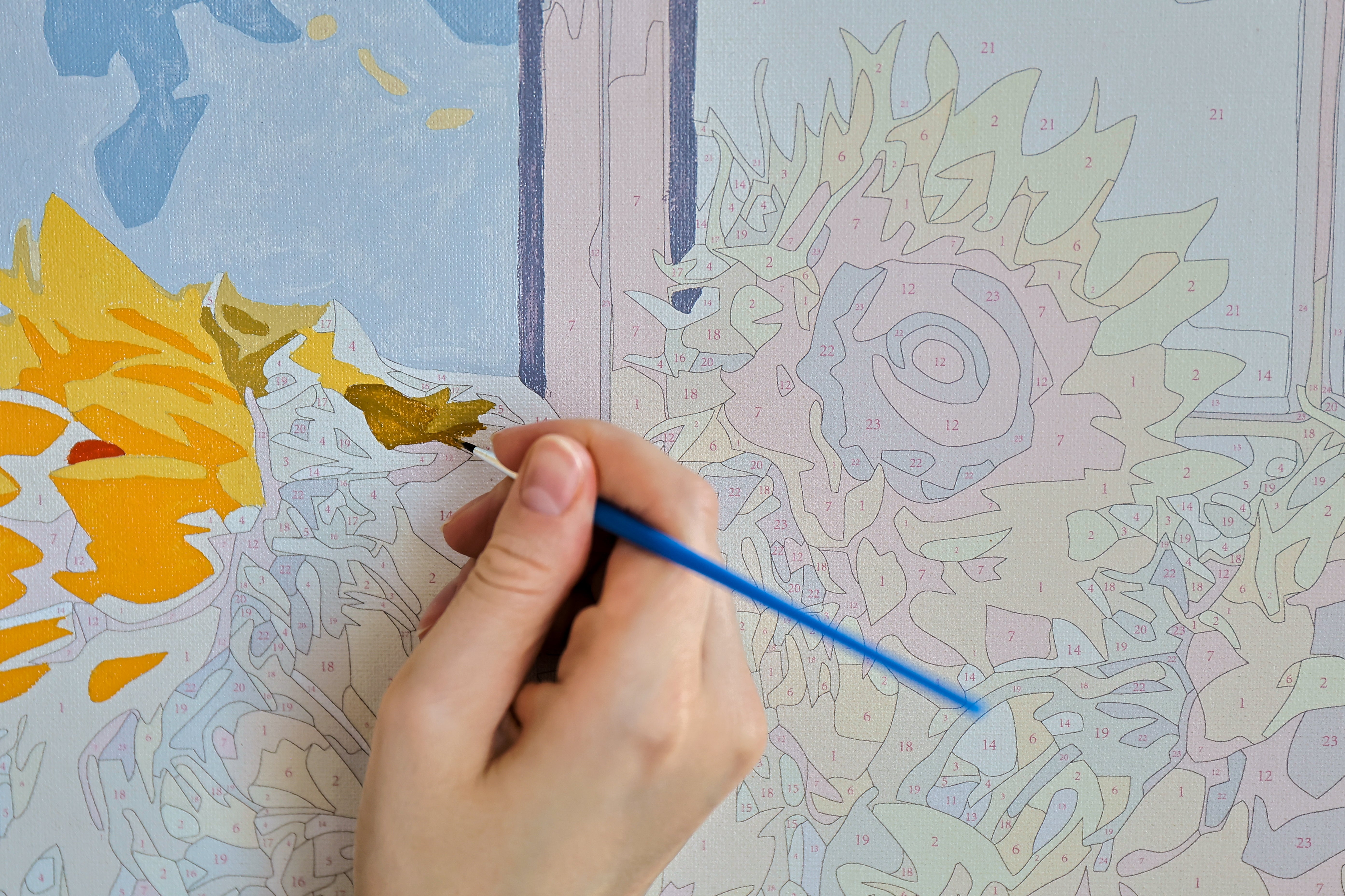 Close-up of a hand using a blue paintbrush on a paint-by-numbers canvas featuring sunflowers