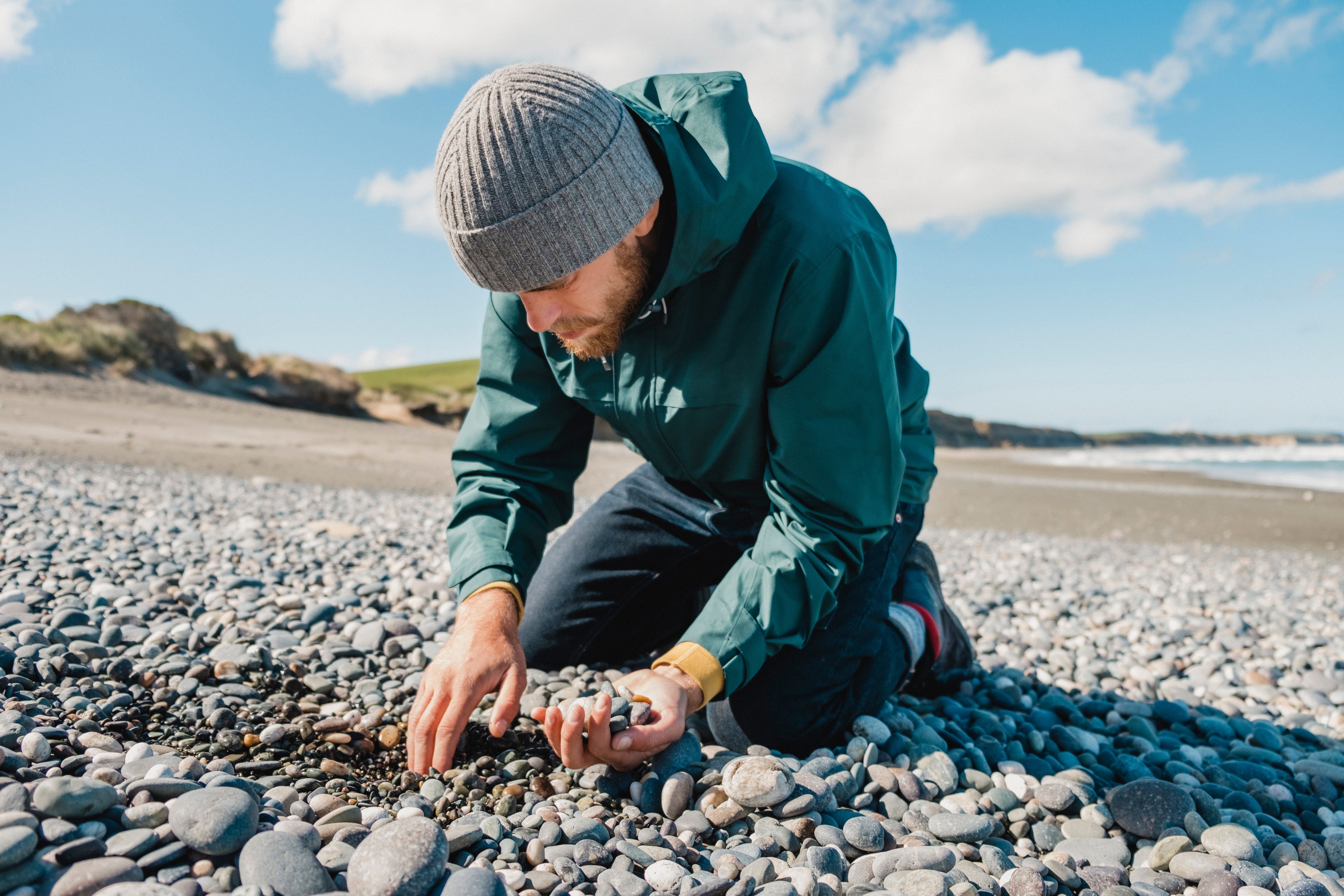 A person in a knit hat and jacket kneels on a rocky beach, examining stones in a peaceful, natural setting