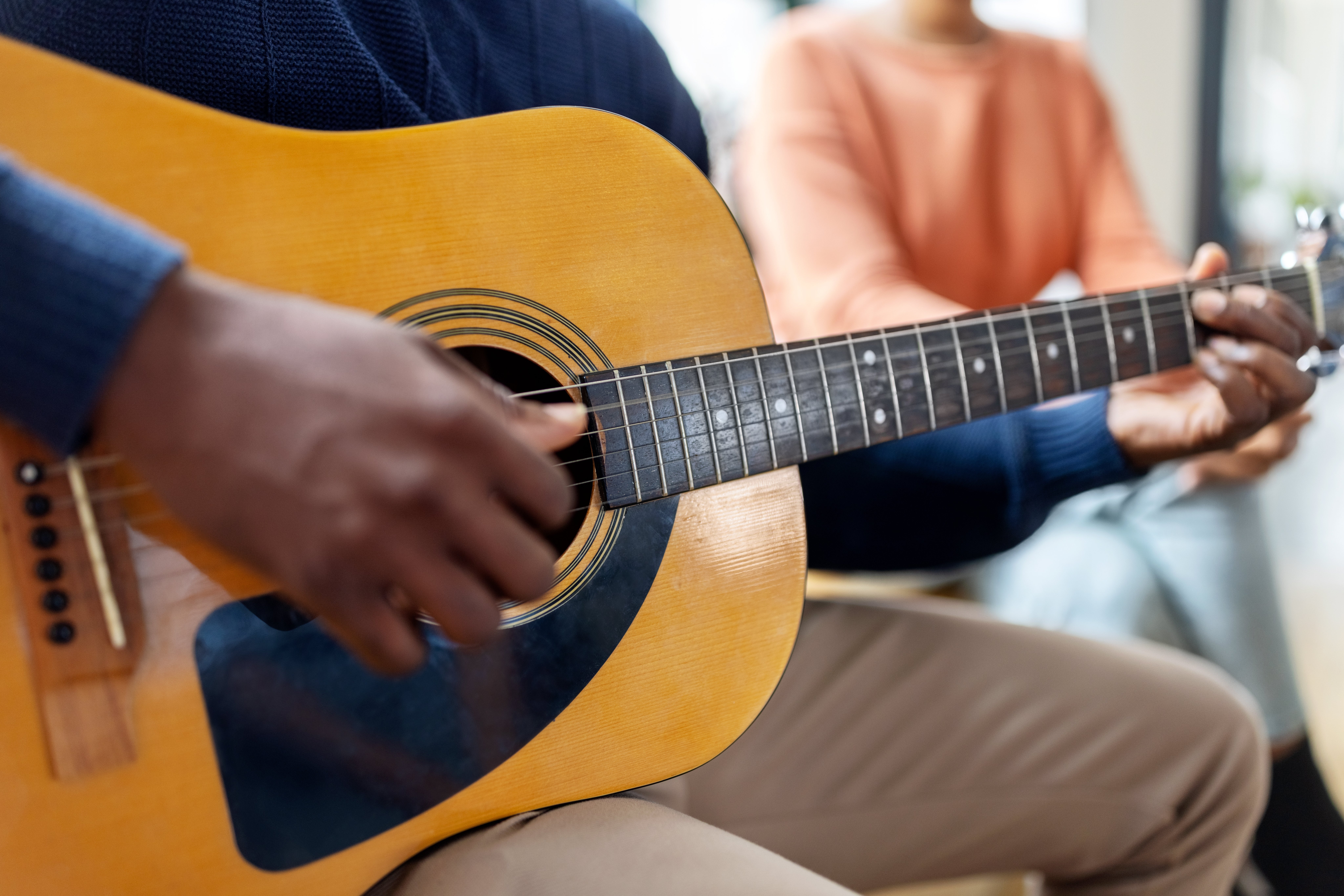 Person playing an acoustic guitar while another person sits beside them, focusing on their hands and the guitar