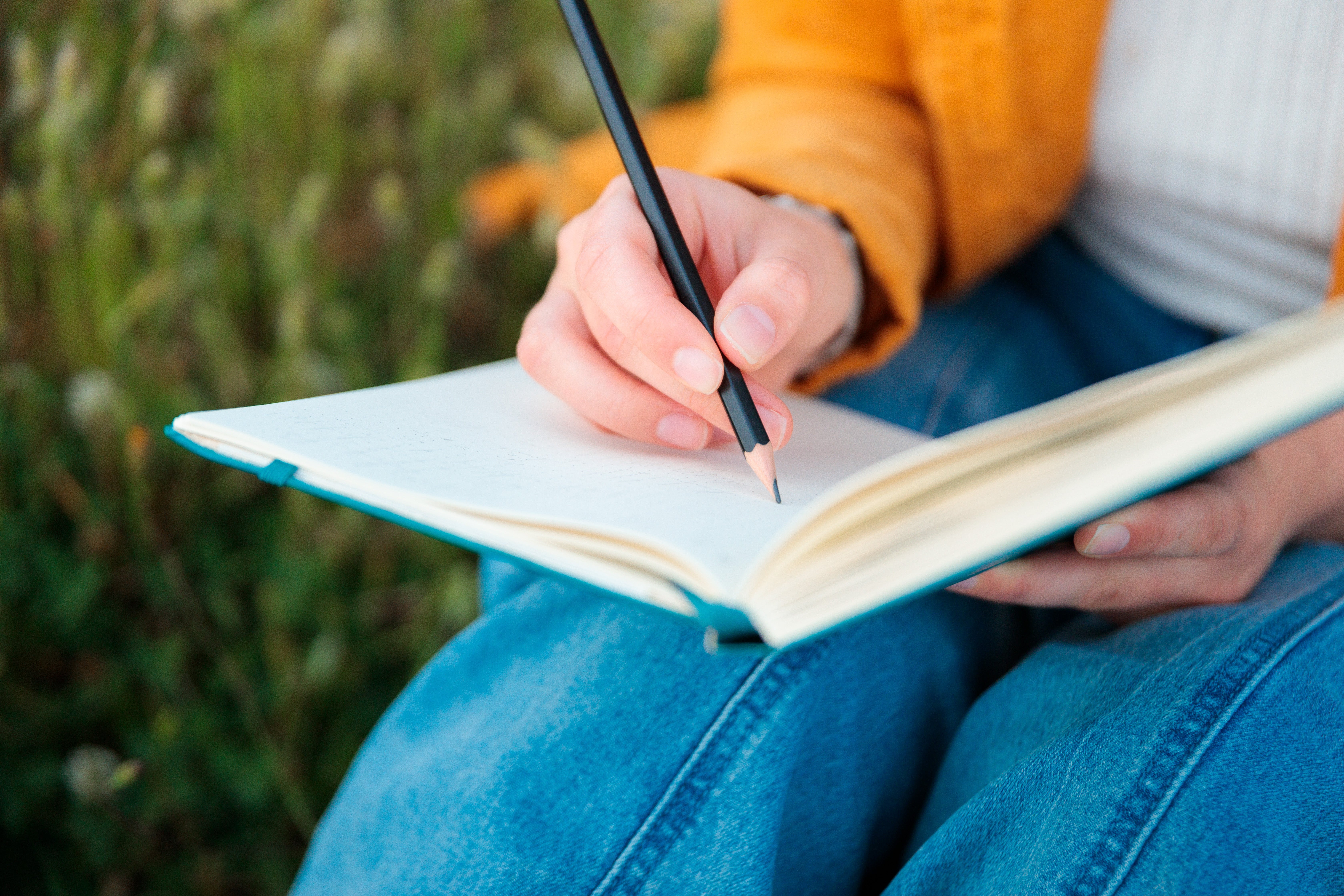 Person writing in a notebook on their lap, surrounded by greenery, suggesting a peaceful outdoor setting