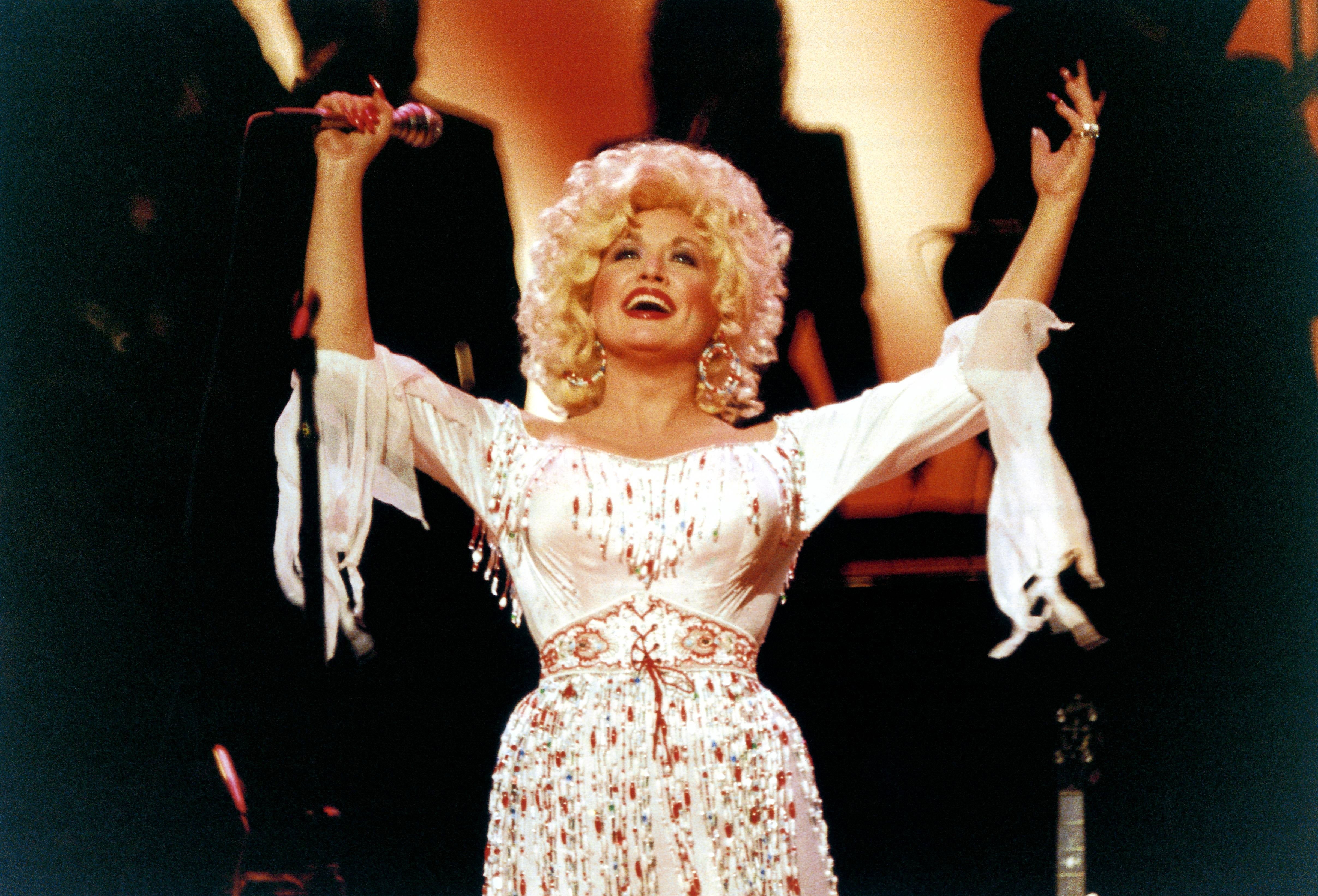 A performer in an embellished, flowing dress raises a microphone and their arms, joyously engaging with the audience on stage