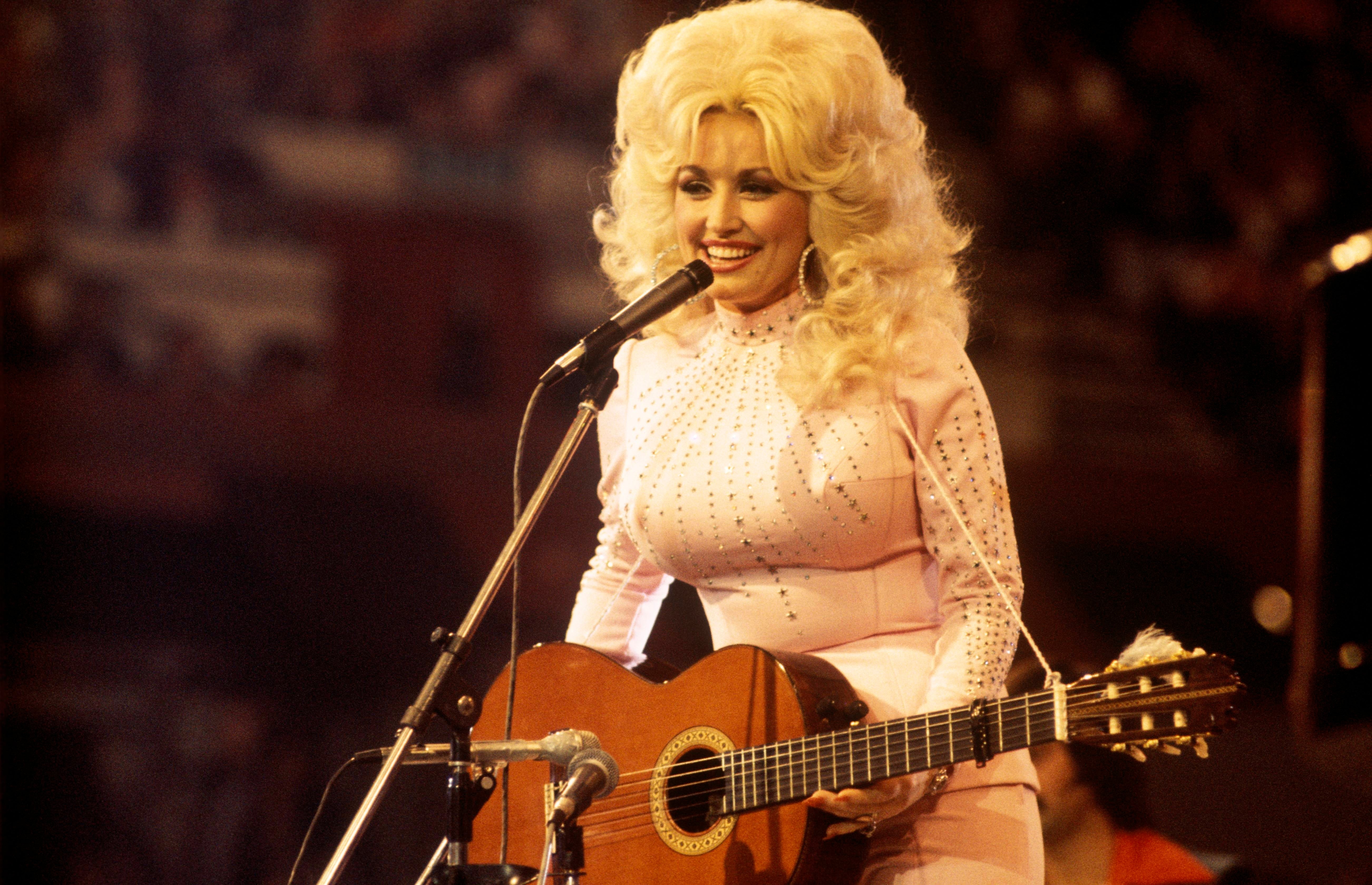 A performer with voluminous hair sings on stage, wearing a sparkling, fitted gown and playing an acoustic guitar