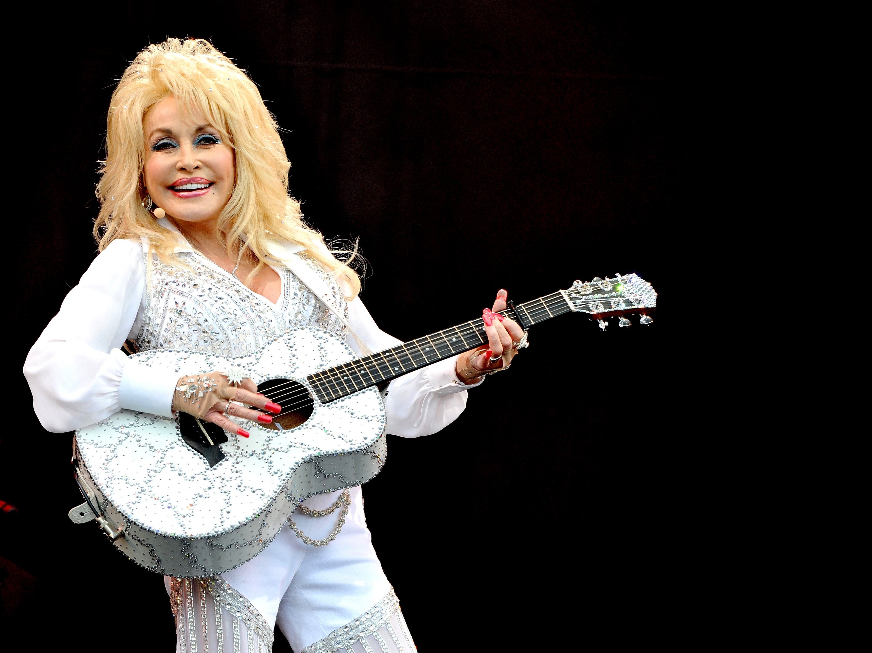 Person playing a rhinestone-studded guitar, wearing a glamorous white outfit with intricate details, smiling on stage