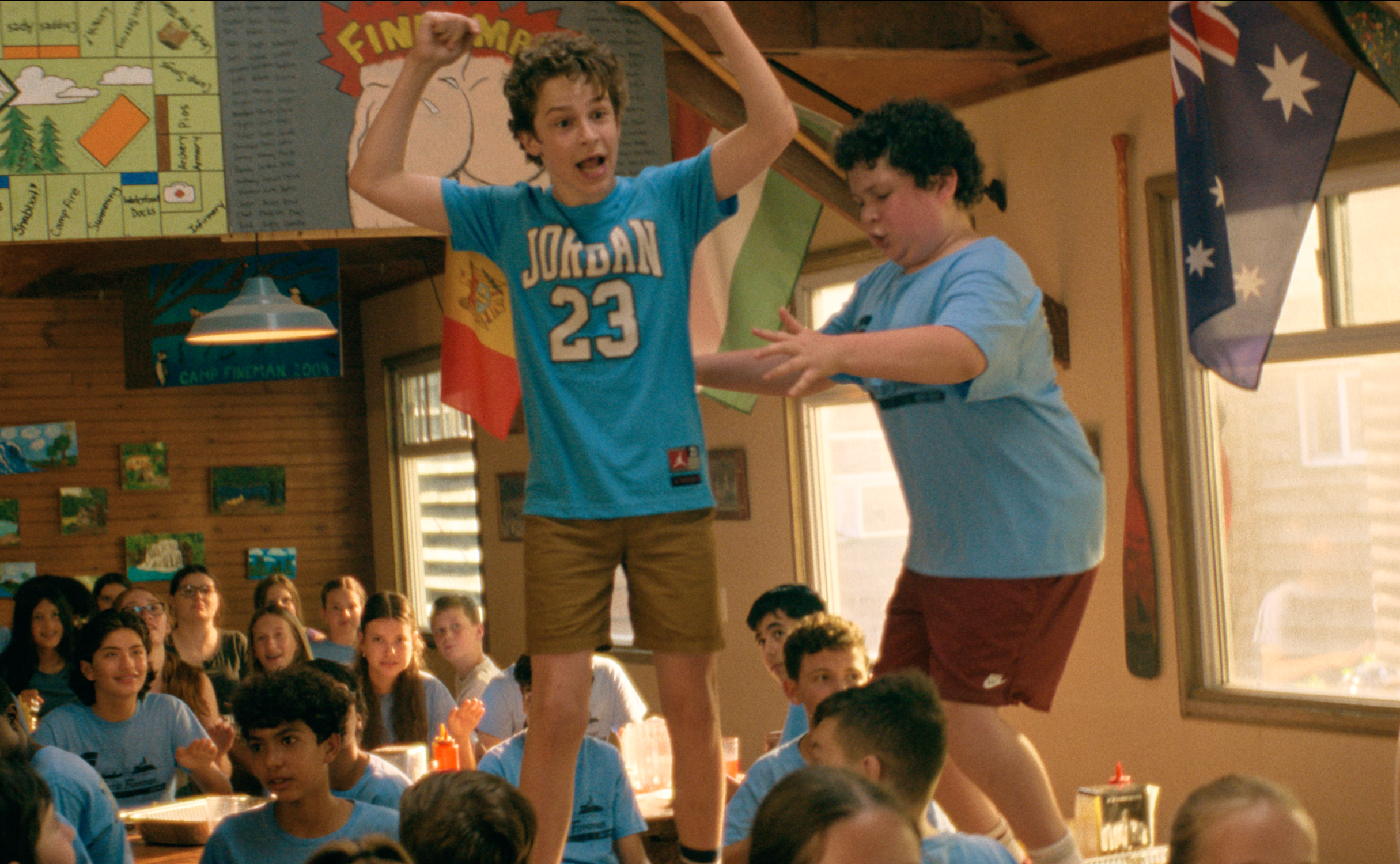 Two kids in a school cafeteria dance on tables, surrounded by classmates. Both wear casual clothes and appear energetic and excited. Flags hang nearby