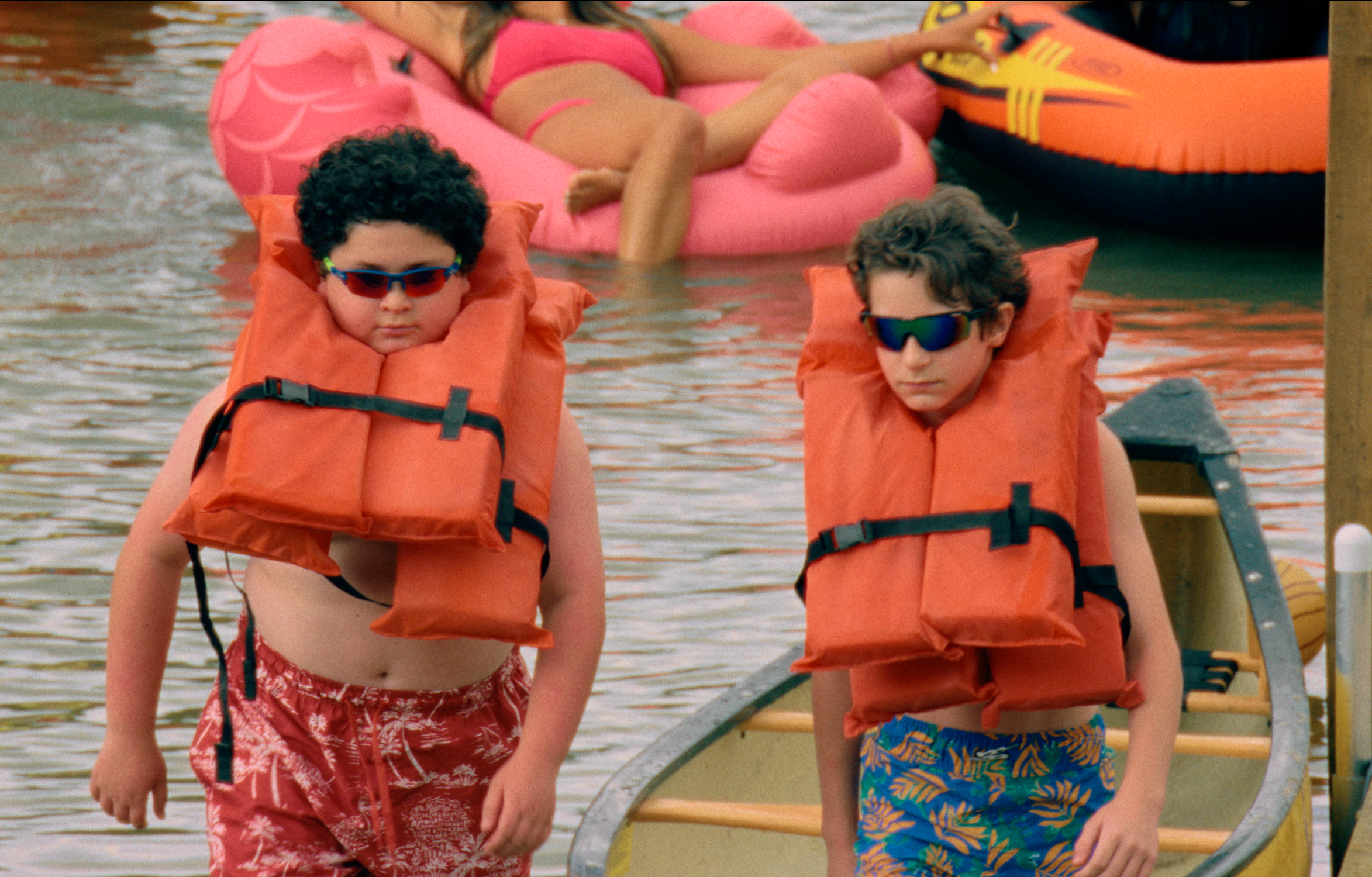 Two kids in life vests and sunglasses walk by water toys. They seem to be at a lakeside or pool area, with a playful summer vibe