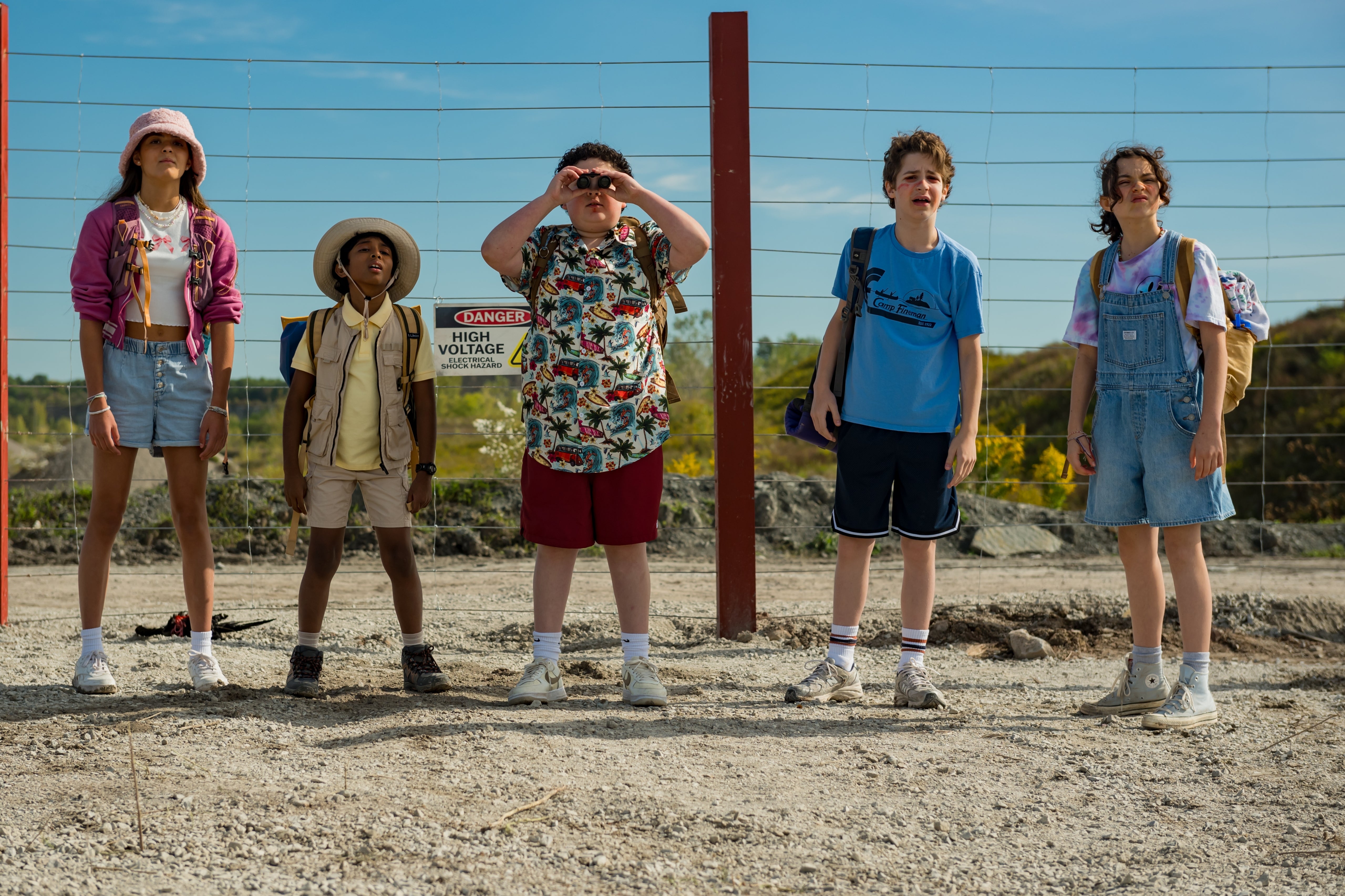Five kids stand on a gravel path near a fence with warning signs, dressed in diverse casual adventure outfits and looking into the distance