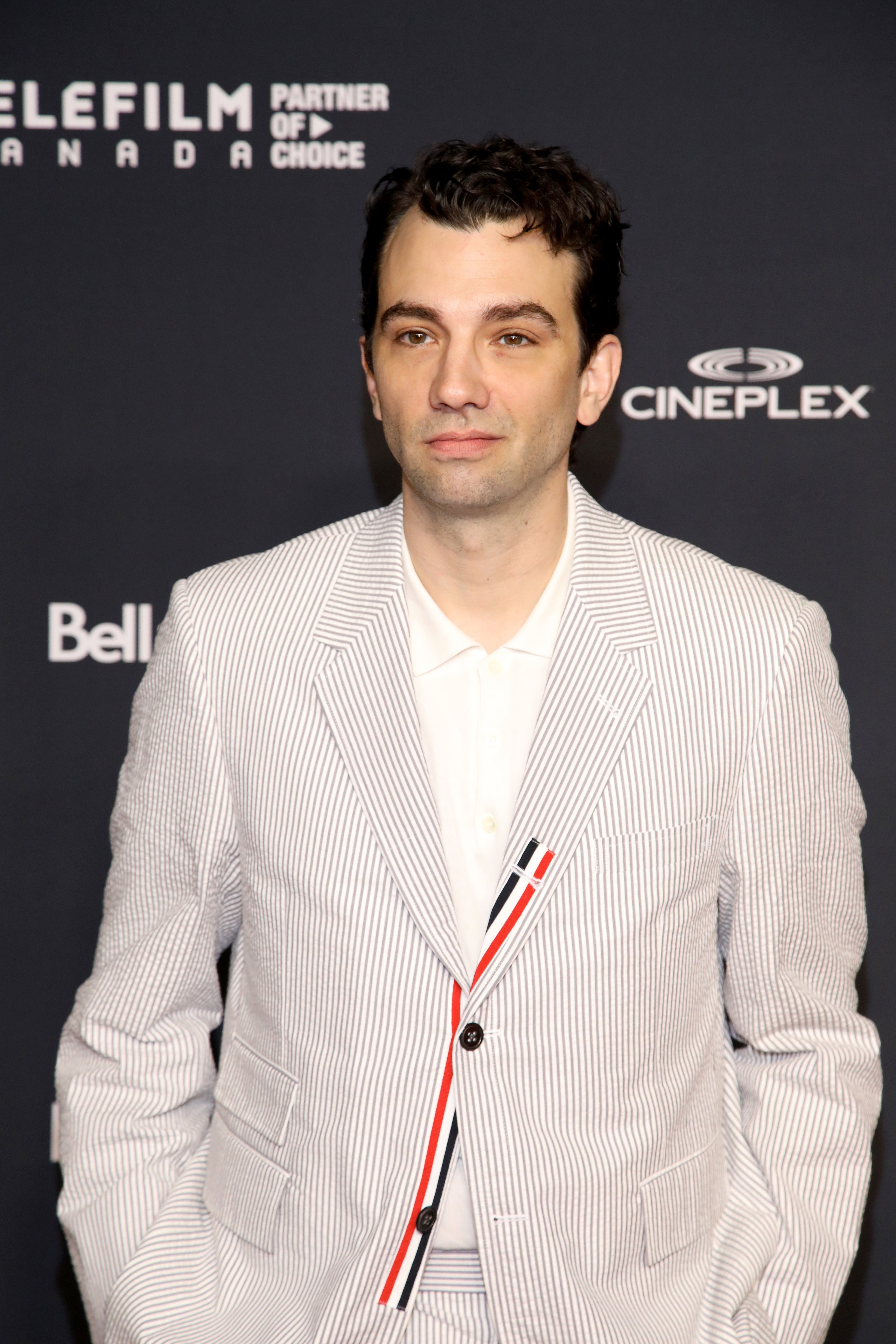 Person on red carpet in a striped suit jacket over a white shirt, standing in front of a backdrop with logos