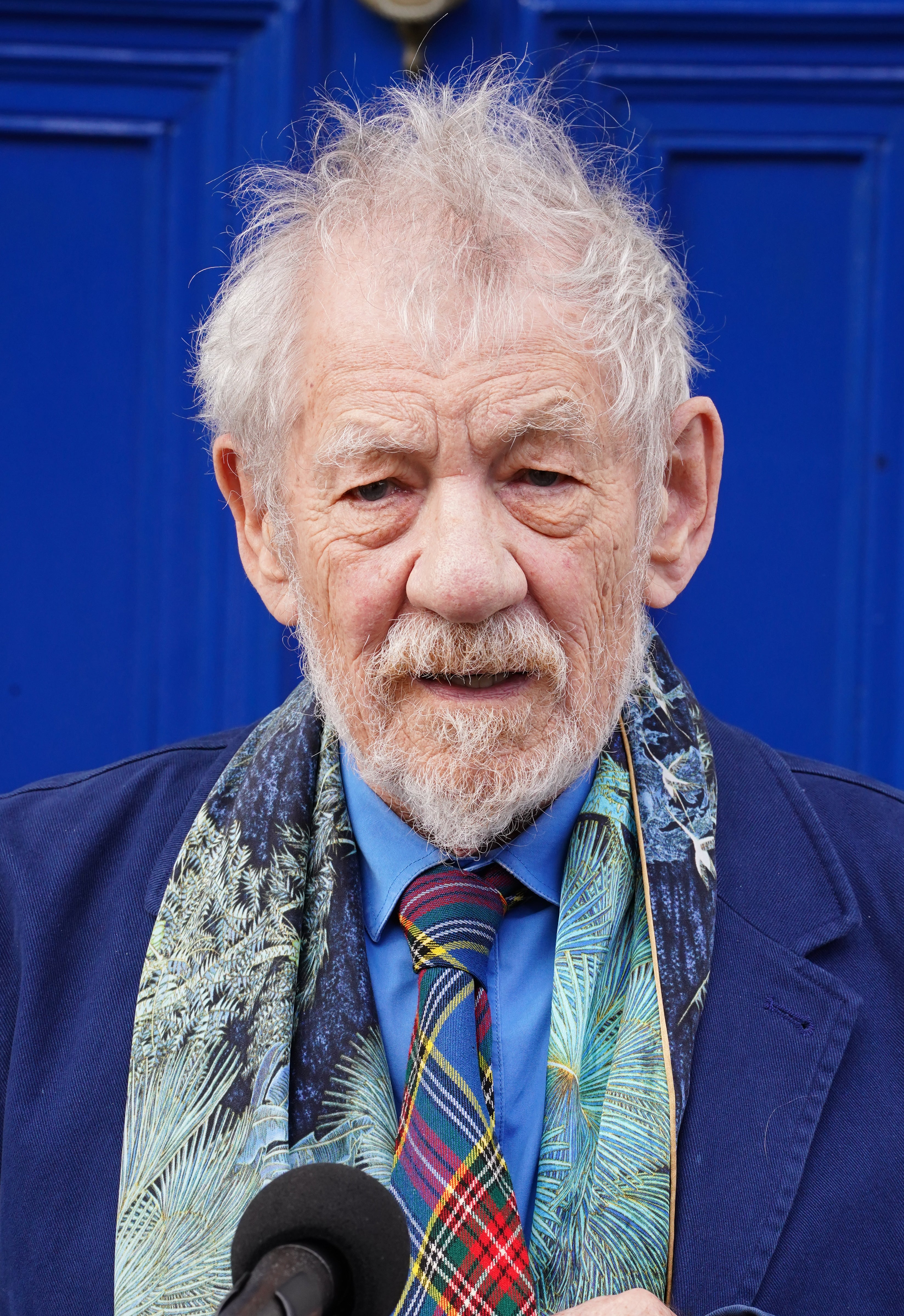 Elderly man in a blue suit with a colorful scarf and tie, standing in front of a door