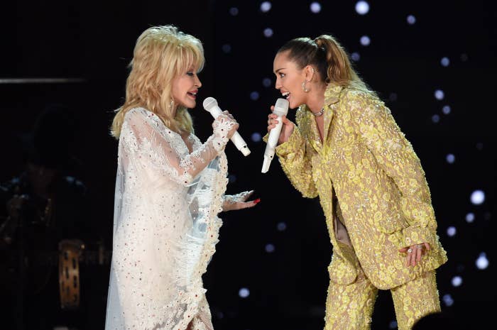 Two women perform on stage. One wears a sparkling white dress, the other a floral-patterned suit. Both hold microphones, facing each other