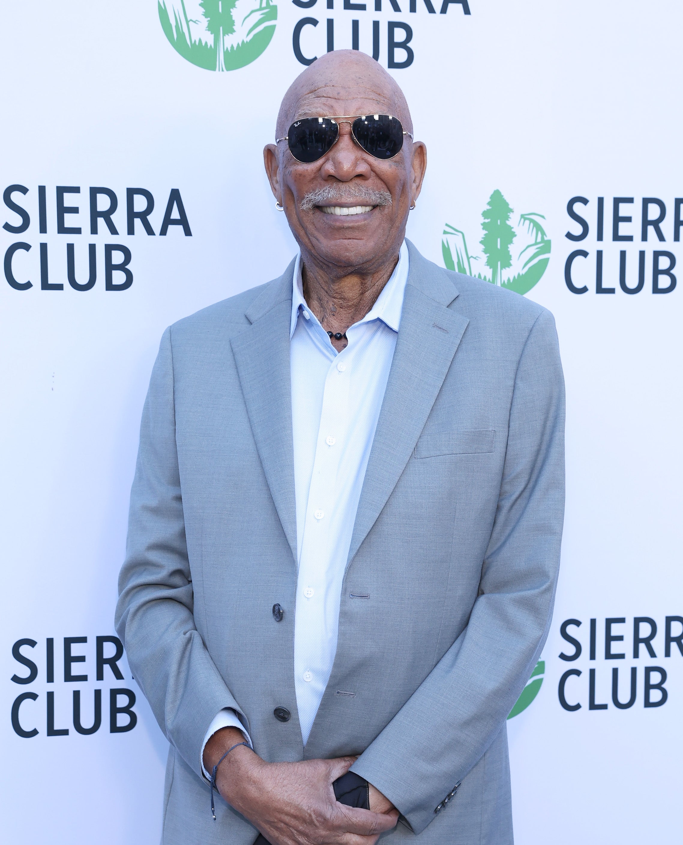 A person in a stylish suit and sunglasses smiles while posing at a Sierra Club event backdrop