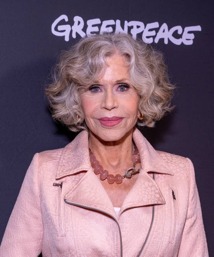 Person with curly hair and a pink jacket poses in front of a backdrop with the Greenpeace logo