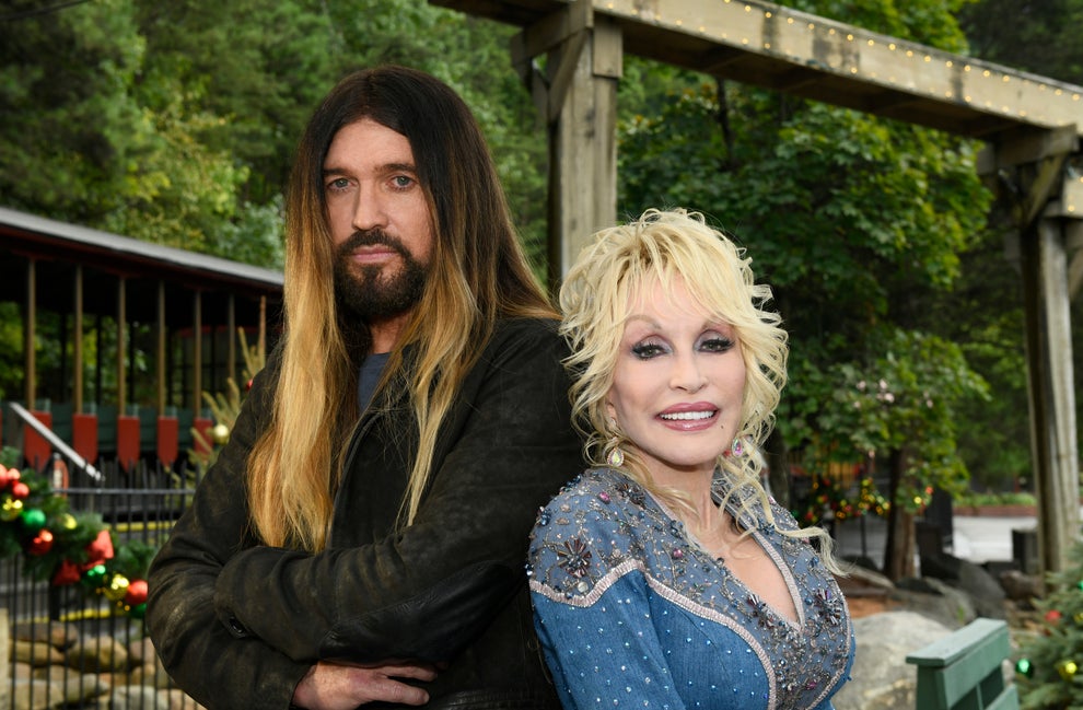 Two celebrities pose together outdoors. The man has long hair and a jacket; the woman wears a denim-style embellished outfit