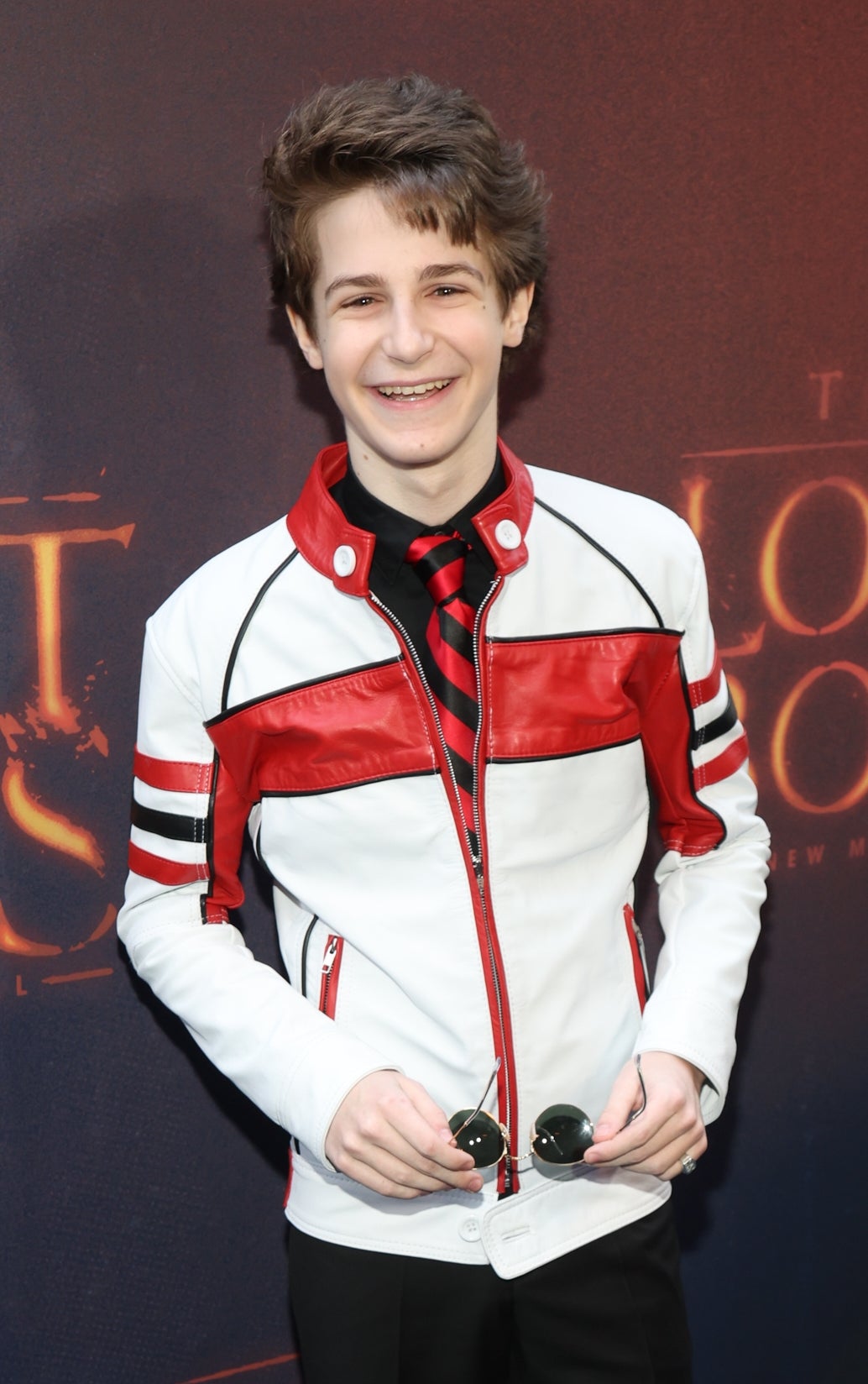 Person in a stylish white leather jacket with red stripes, holding sunglasses, at "The Lost Boys" musical event