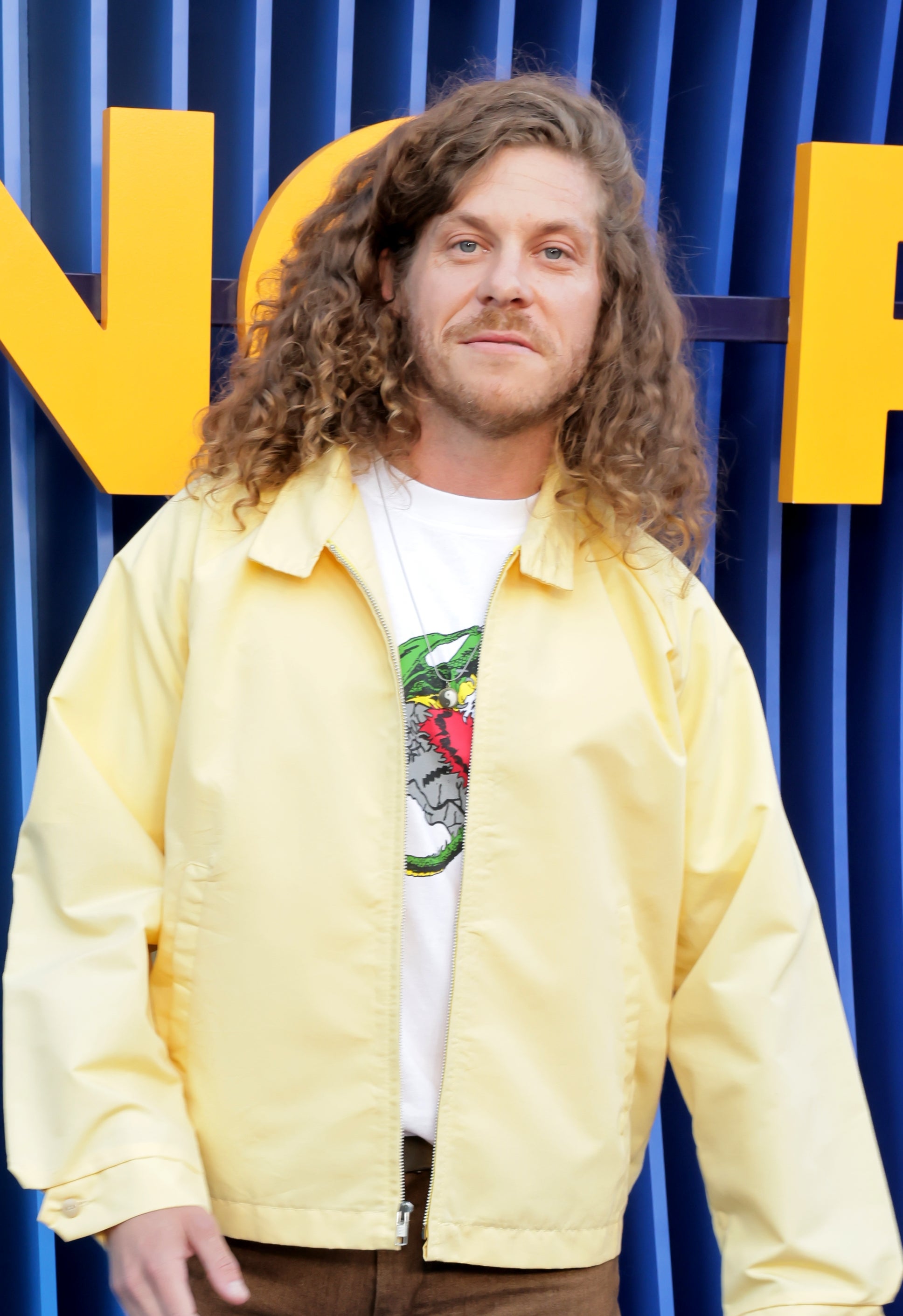Person with long hair in casual attire: yellow jacket, graphic tee, brown pants, and sneakers, posing at an event with a patterned backdrop
