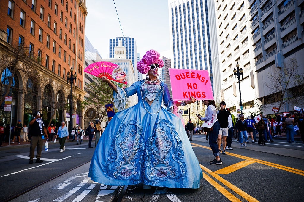Person in elaborate historical costume and wig holds fans and sign reading "Queens, No Kings" at a street event with city buildings in background