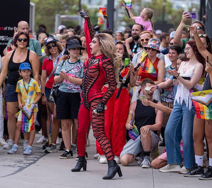 A drag performer in a striking, form-fitting red and black outfit poses confidently with a diverse, cheering crowd holding Pride flags