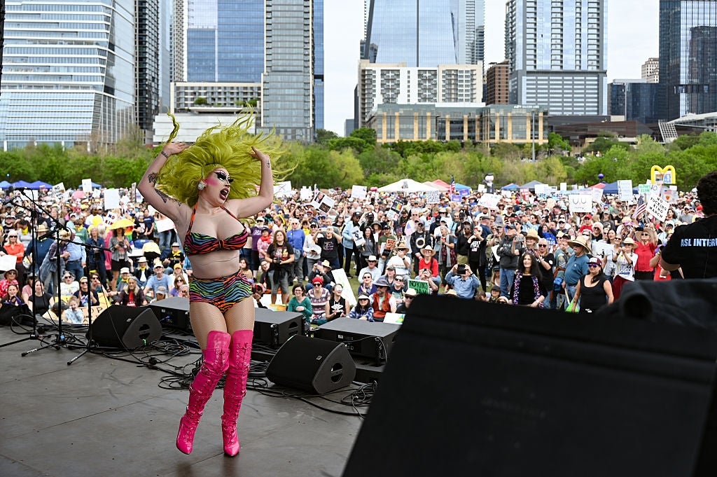 Performer wearing a vibrant outfit and striking boots dances energetically on stage during an outdoor LGBT event with a large crowd in attendance