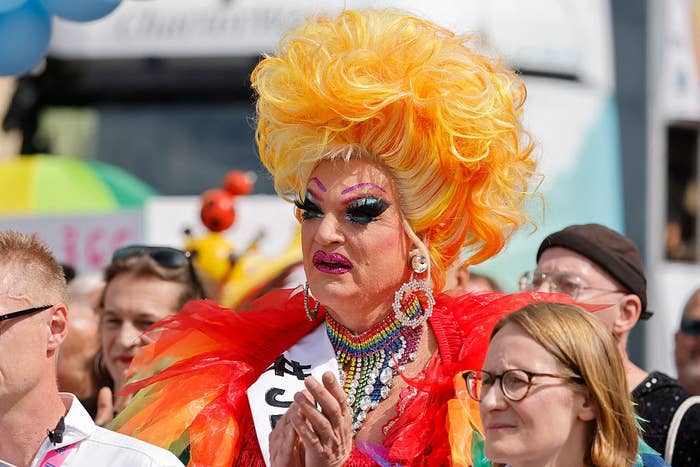 Person in vibrant drag attire with voluminous hair, colorful feathers, and bold makeup, clapping in a lively crowd at an outdoor event
