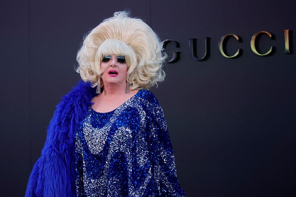 Person in an extravagant sequined dress and large wig on red carpet in front of Gucci sign