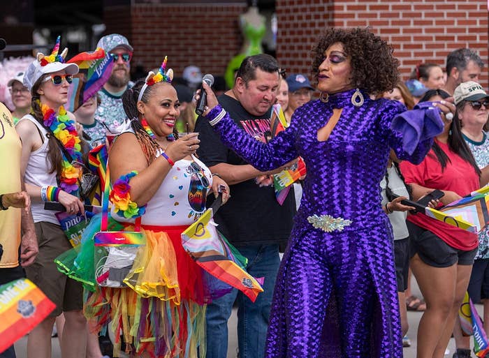 Two people in vibrant, rainbow-themed outfits celebrate at a Pride event, surrounded by a cheerful crowd holding Pride flags