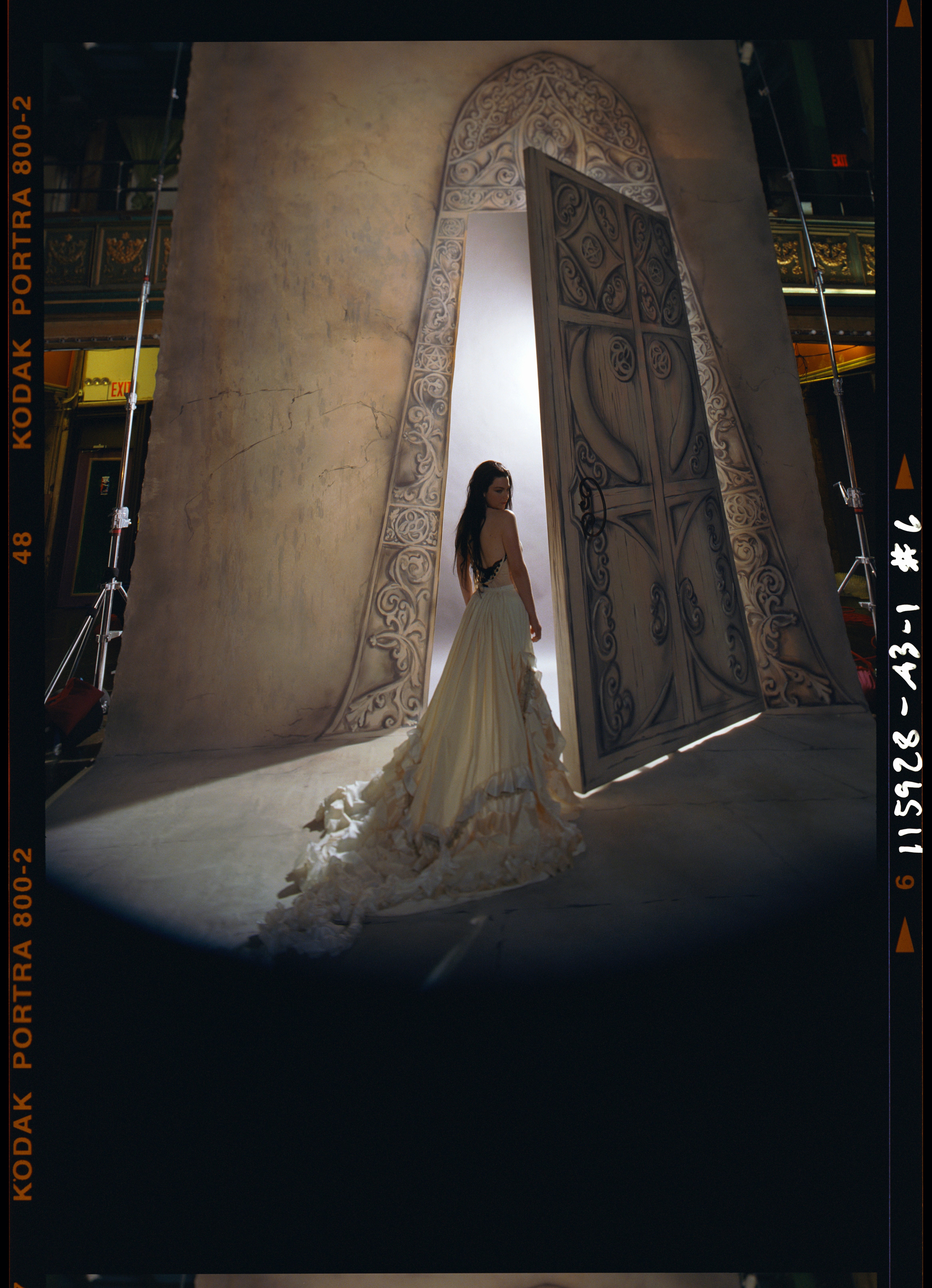 A person in an elegant gown stands dramatically near a large, ornate door under stage lighting, possibly at a music event
