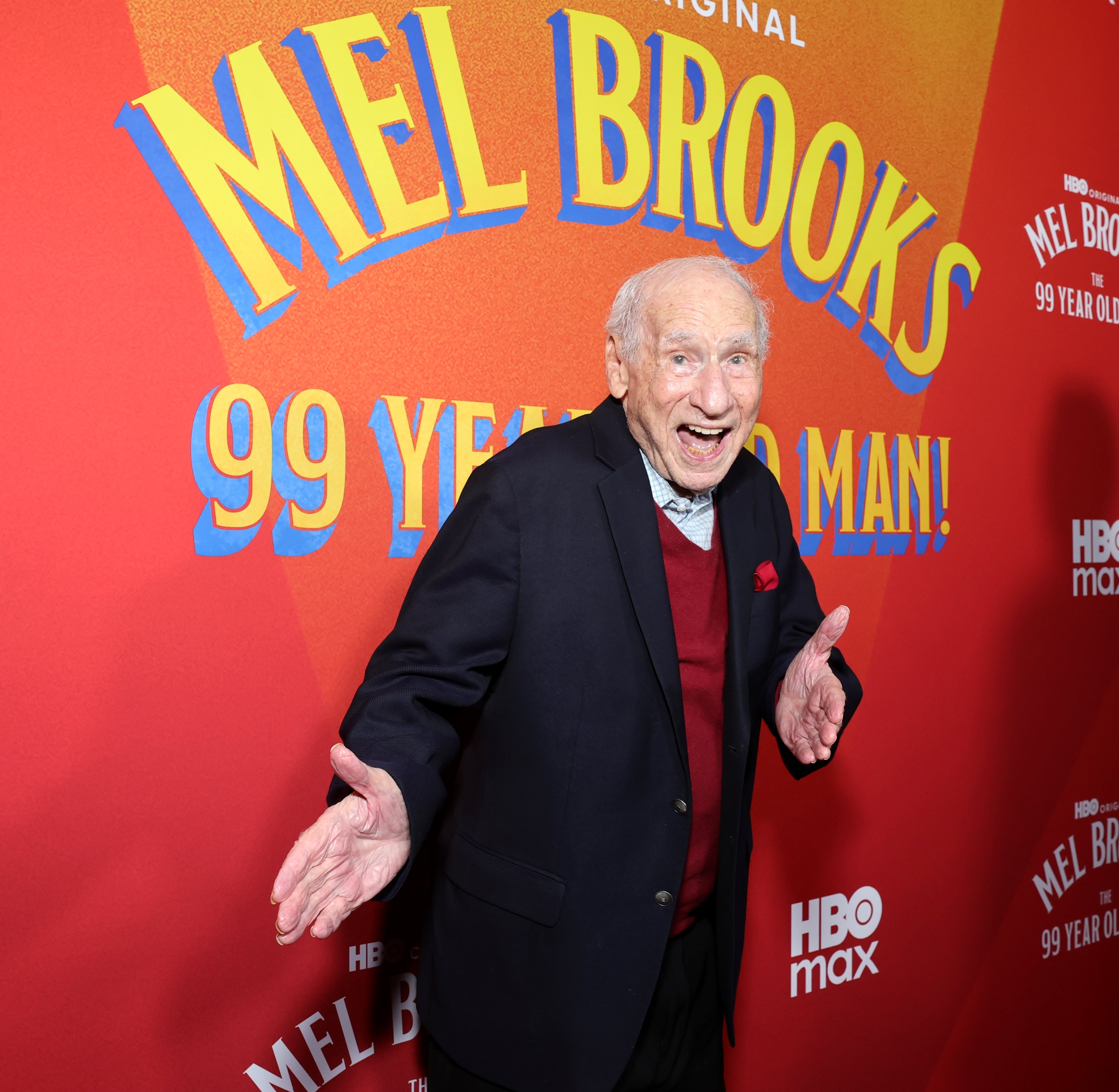 Person on a red carpet event, wearing a blazer over a sweater, smiling and posing in front of a backdrop celebrating Mel Brooks’ 99 years