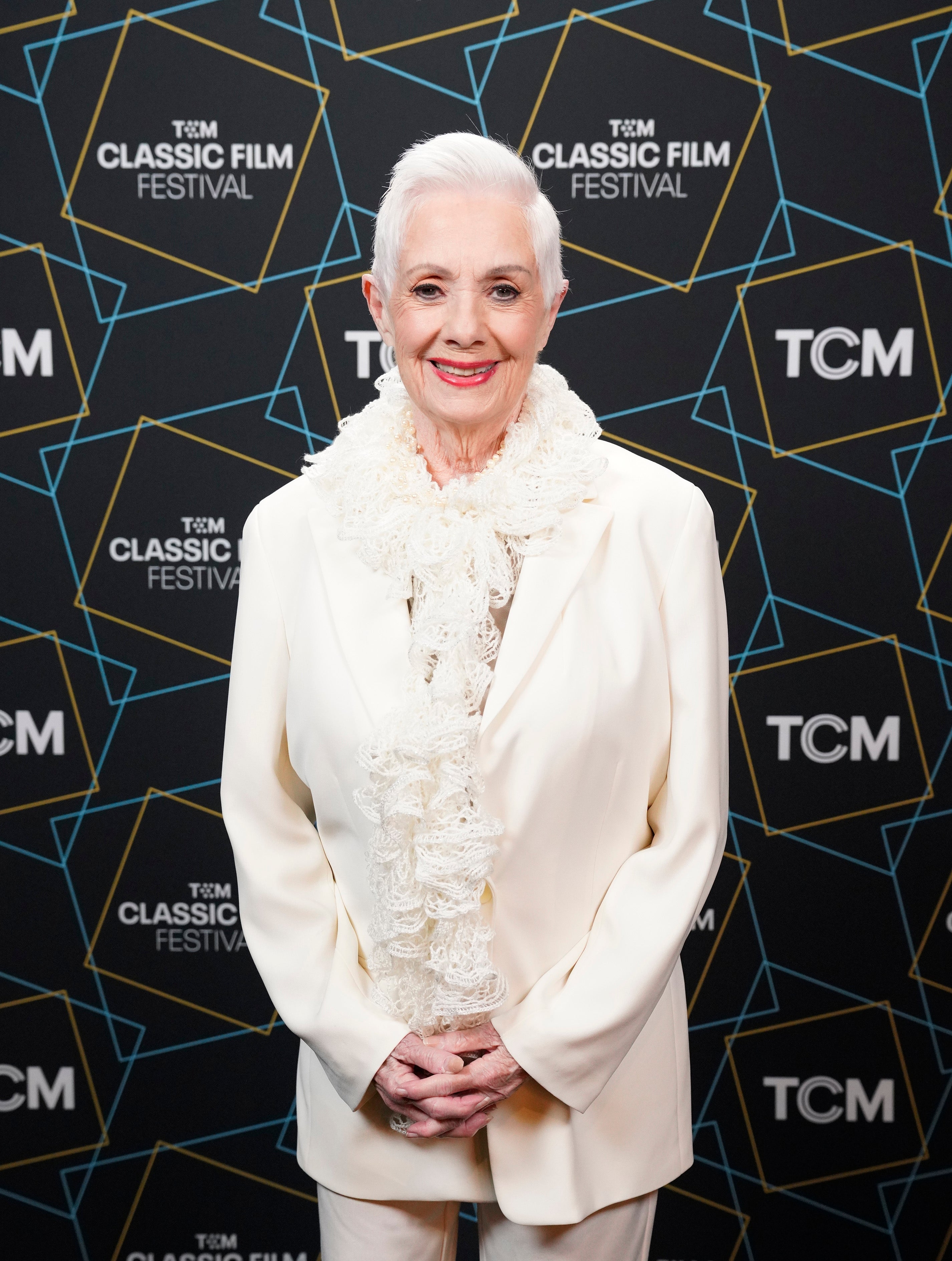 Person smiling at a film festival, wearing an elegant white suit with a floral scarf, standing against a TCM Classic Film Festival backdrop