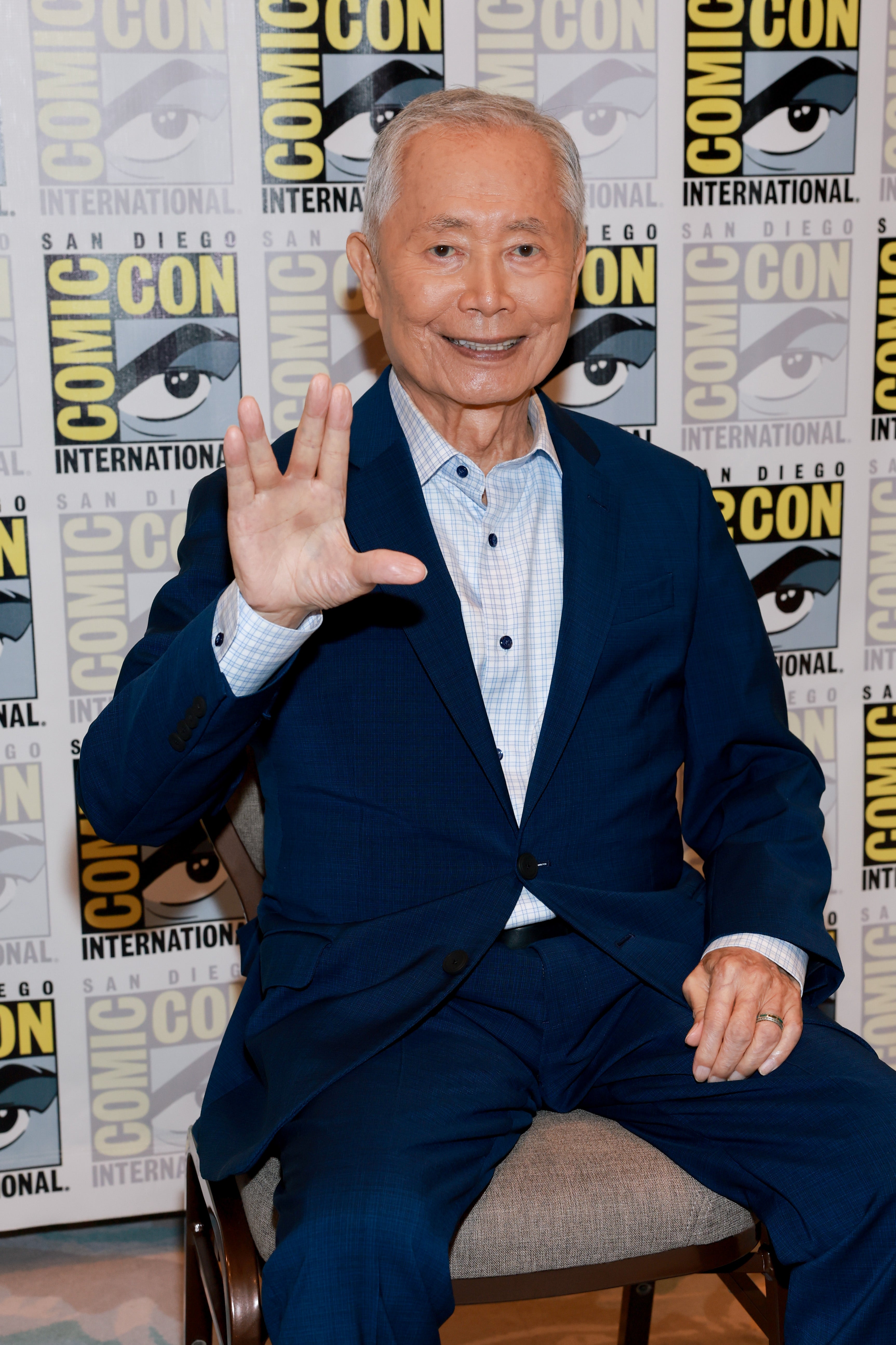 Person at Comic-Con smiling and holding up a Vulcan salute, seated in front of a Comic-Con backdrop, wearing a stylish suit