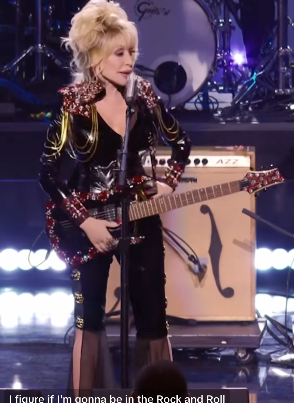 Person in a black, embellished outfit playing a guitar on stage with band equipment in the background. Subtitles mention Rock and Roll