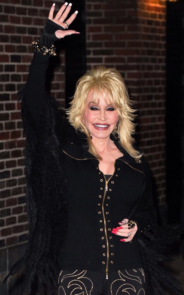 Person smiling and waving, wearing a black outfit with gold accents and decorative detailing, posing near a brick wall