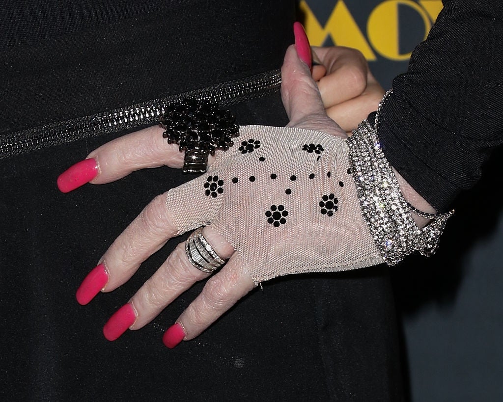 Close-up of hands with mesh gloves, pink nails, black and silver rings, and sparkling bracelets. Female in a black outfit poses with hands on hips
