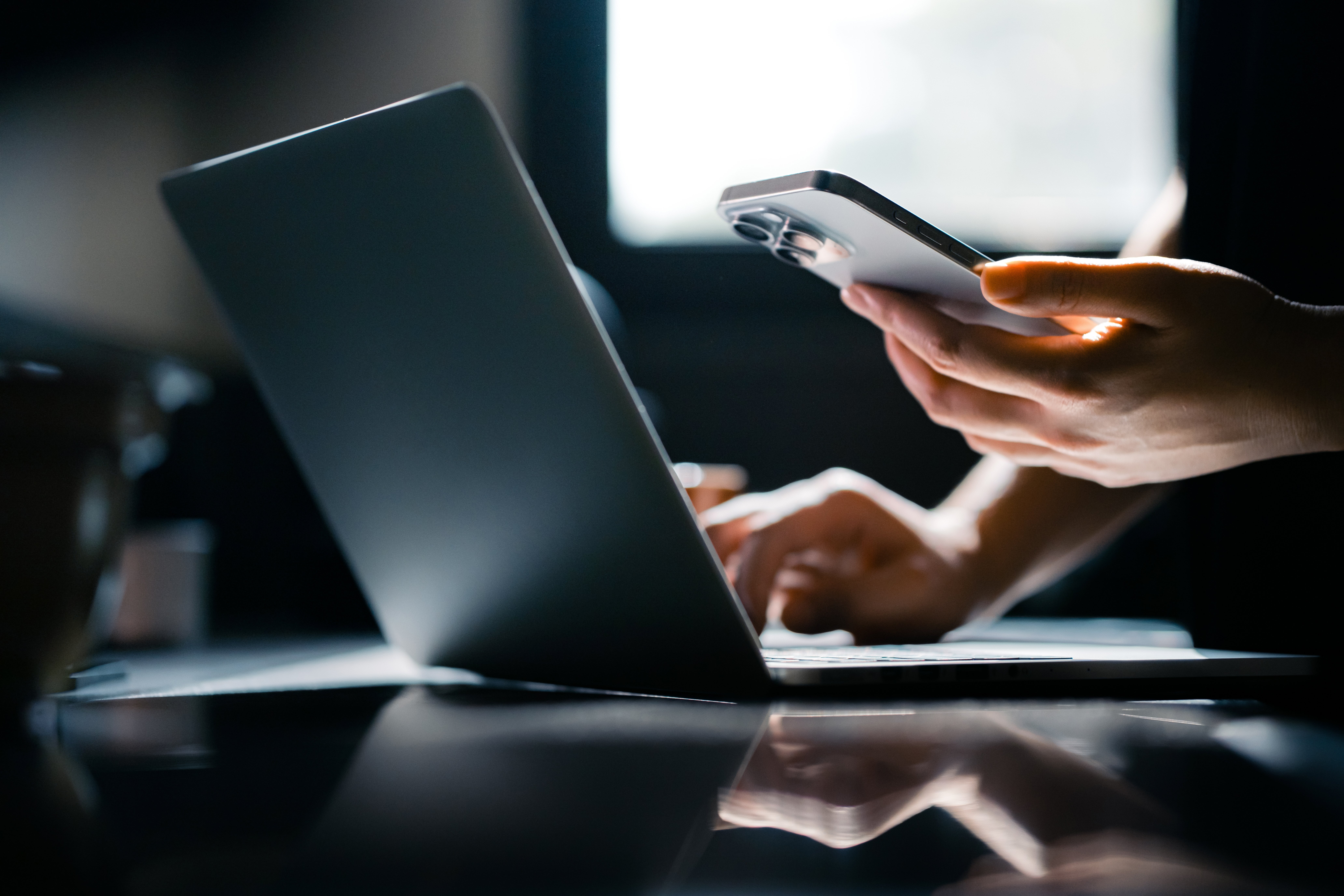 Person using a smartphone and laptop at a desk, suggesting multitasking or remote work setup