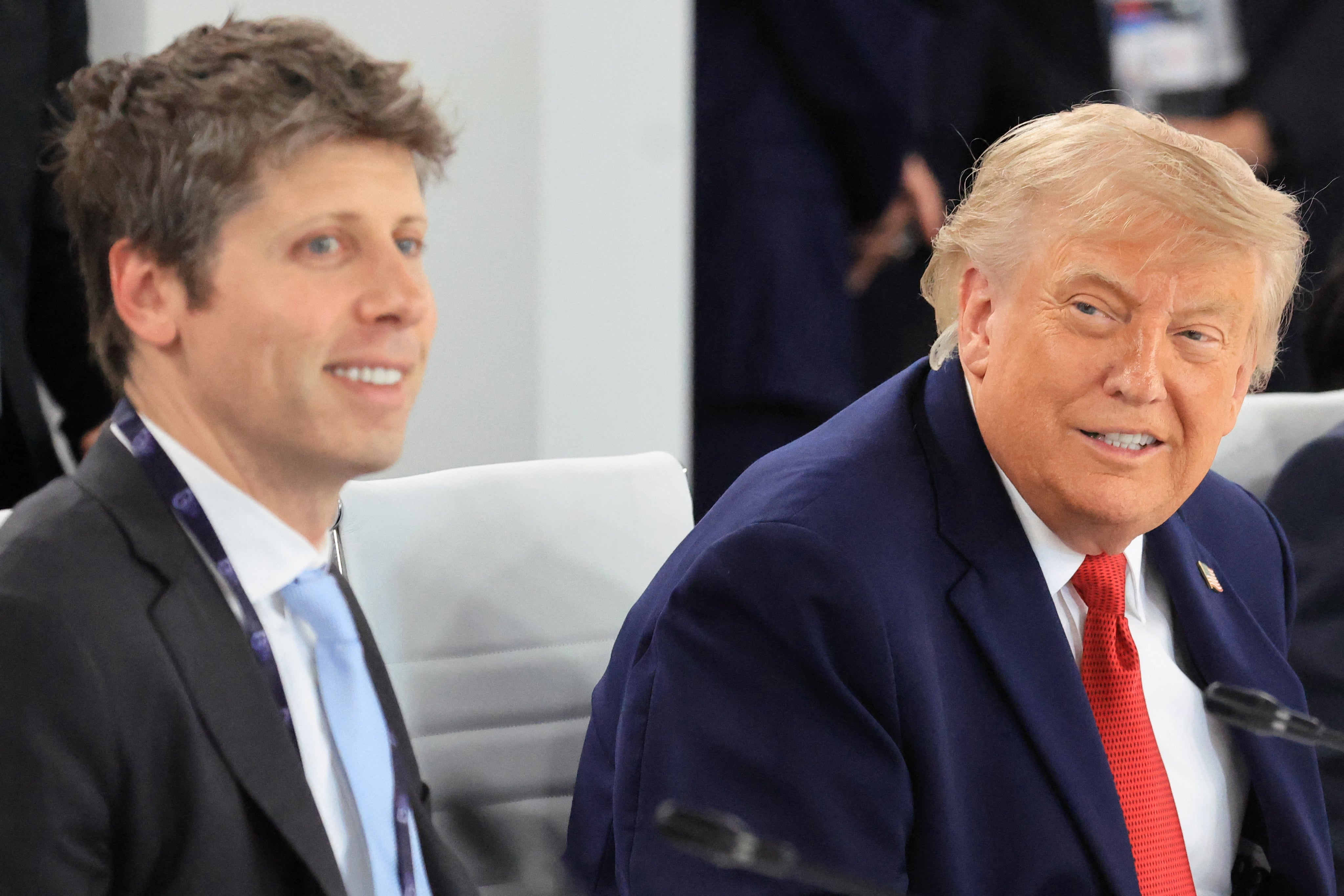 Two men in suits sit at a conference; one smiles while turning toward the other, who is speaking