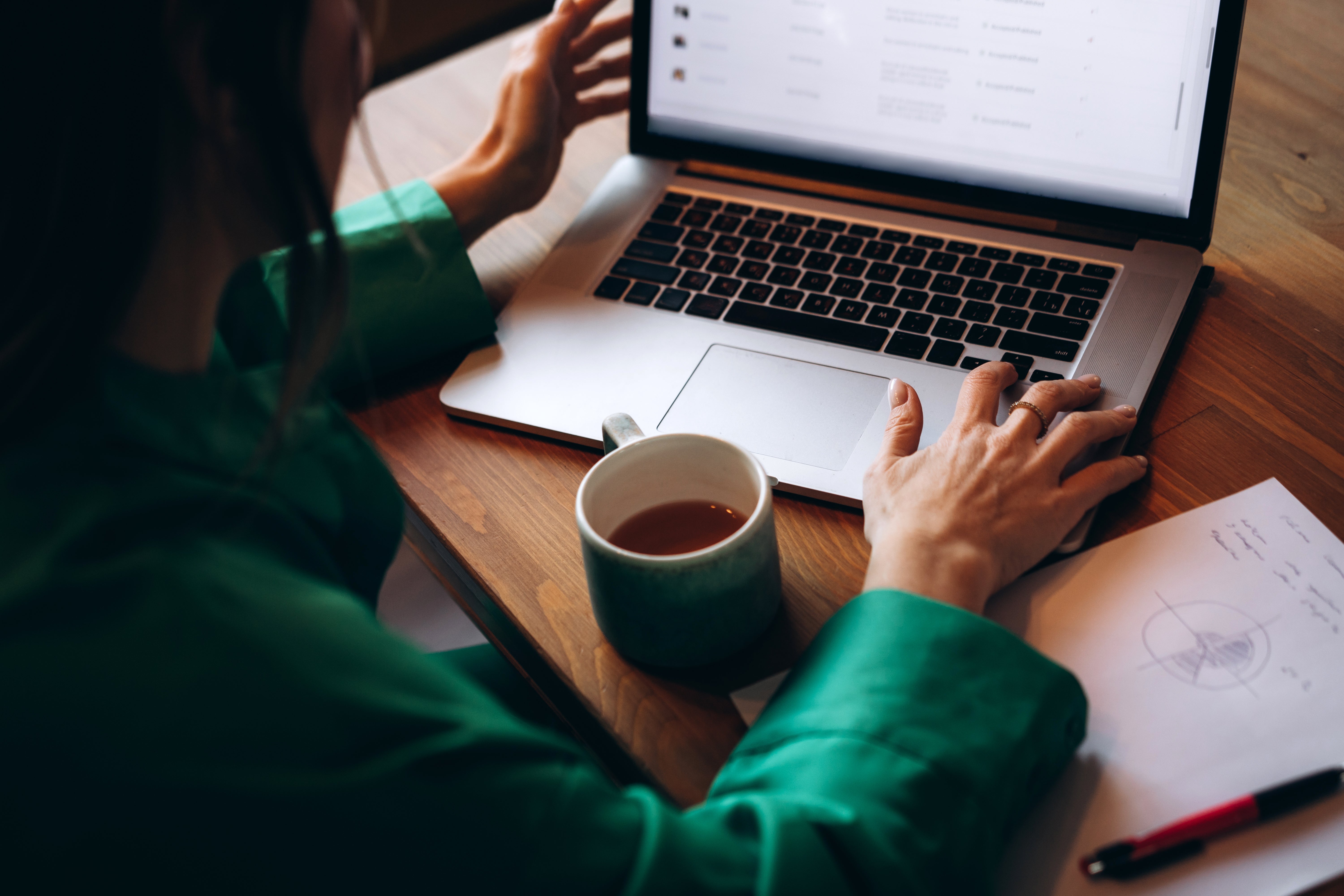 Person in green shirt working on a laptop at a wooden table with coffee and notes