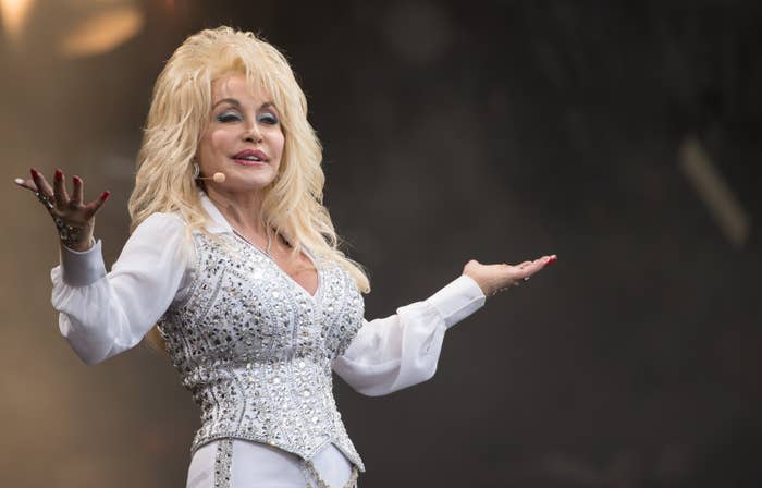 A person in a sparkling, embellished white outfit gestures with open arms on stage, likely during a performance or event