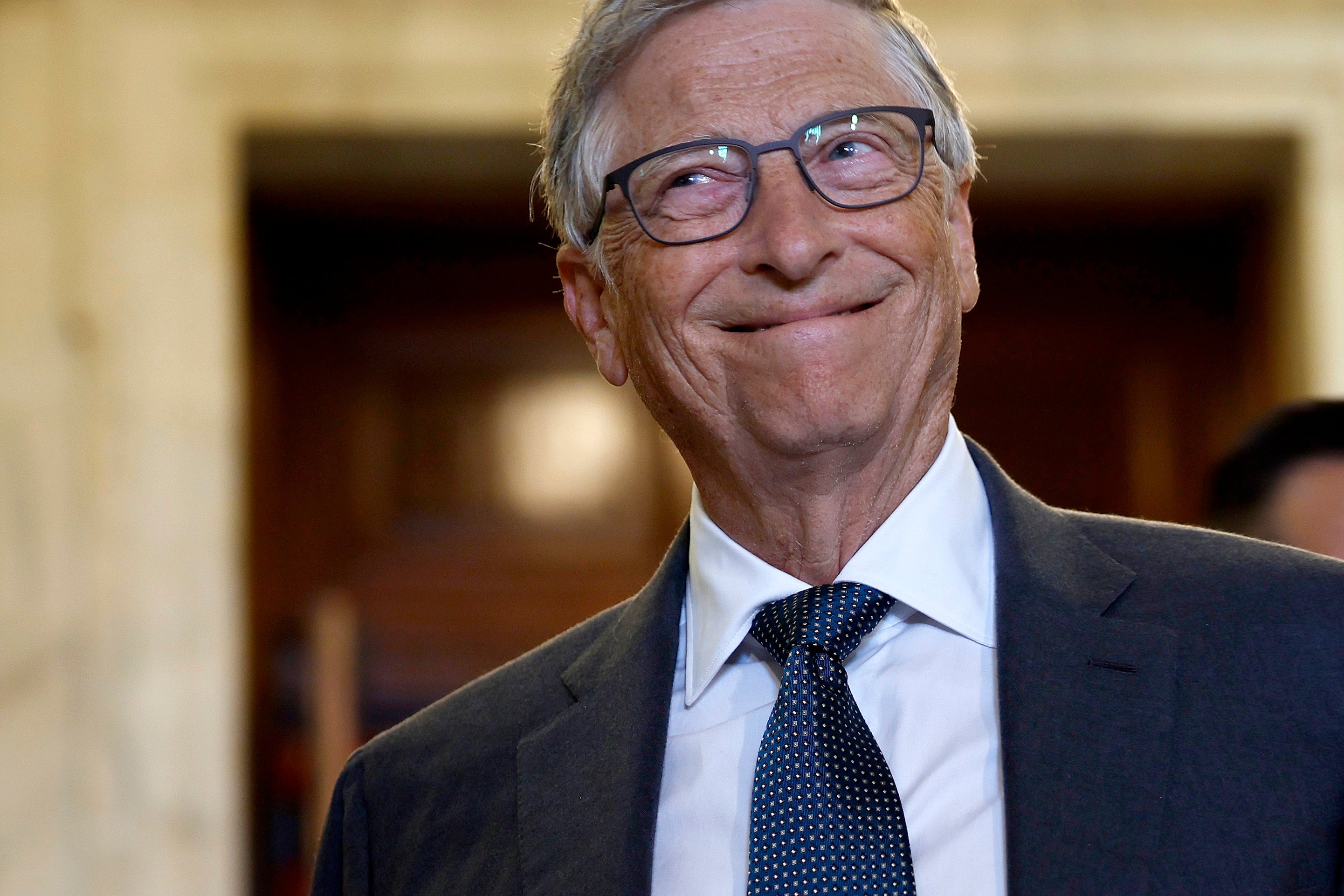 A person in a formal suit and tie smiles while looking off to the side in an indoor setting