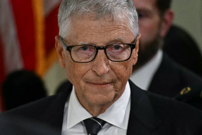 An individual in formal attire, wearing glasses, is standing in front of a blurred background possibly at an event or gathering