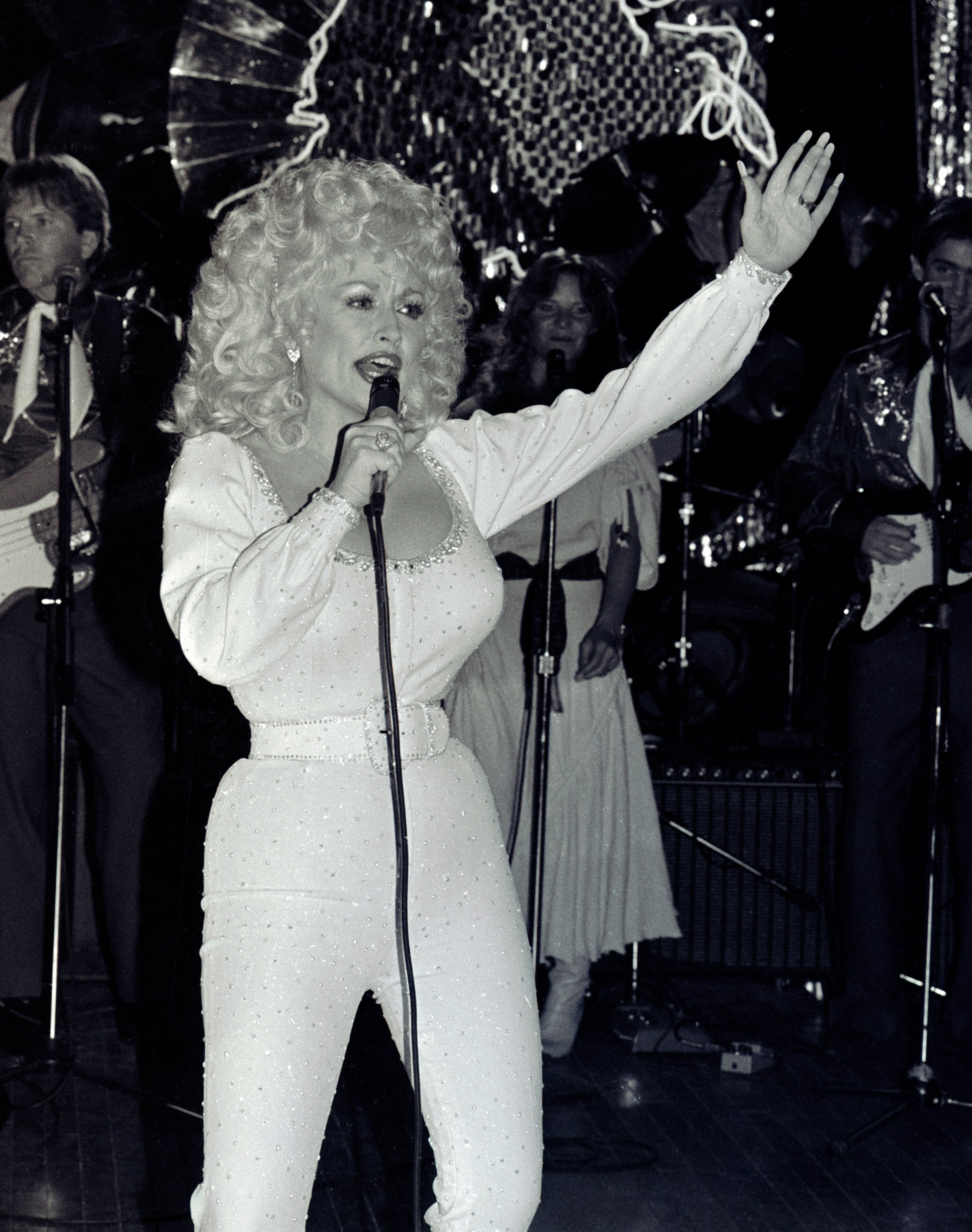 Singer performing on stage with a microphone, wearing a sparkling jumpsuit and surrounded by a band
