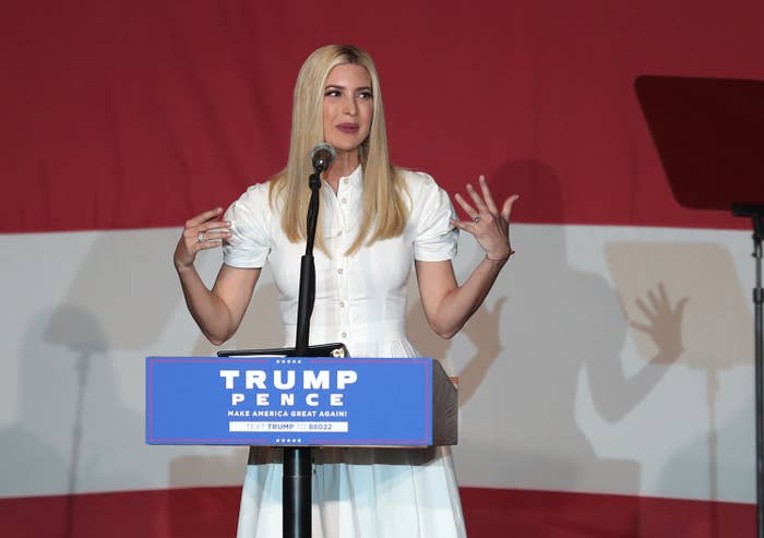A woman is speaking at a podium with a microphone, wearing a short-sleeve, collared dress. There's a red and white backdrop behind her
