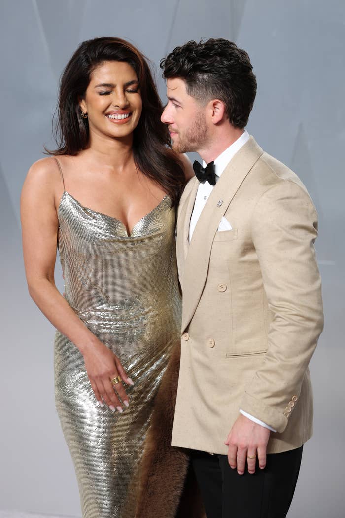 Two people on a red carpet; one wearing a shimmering gown and the other in a beige suit with a bow tie, both smiling and engaged in conversation