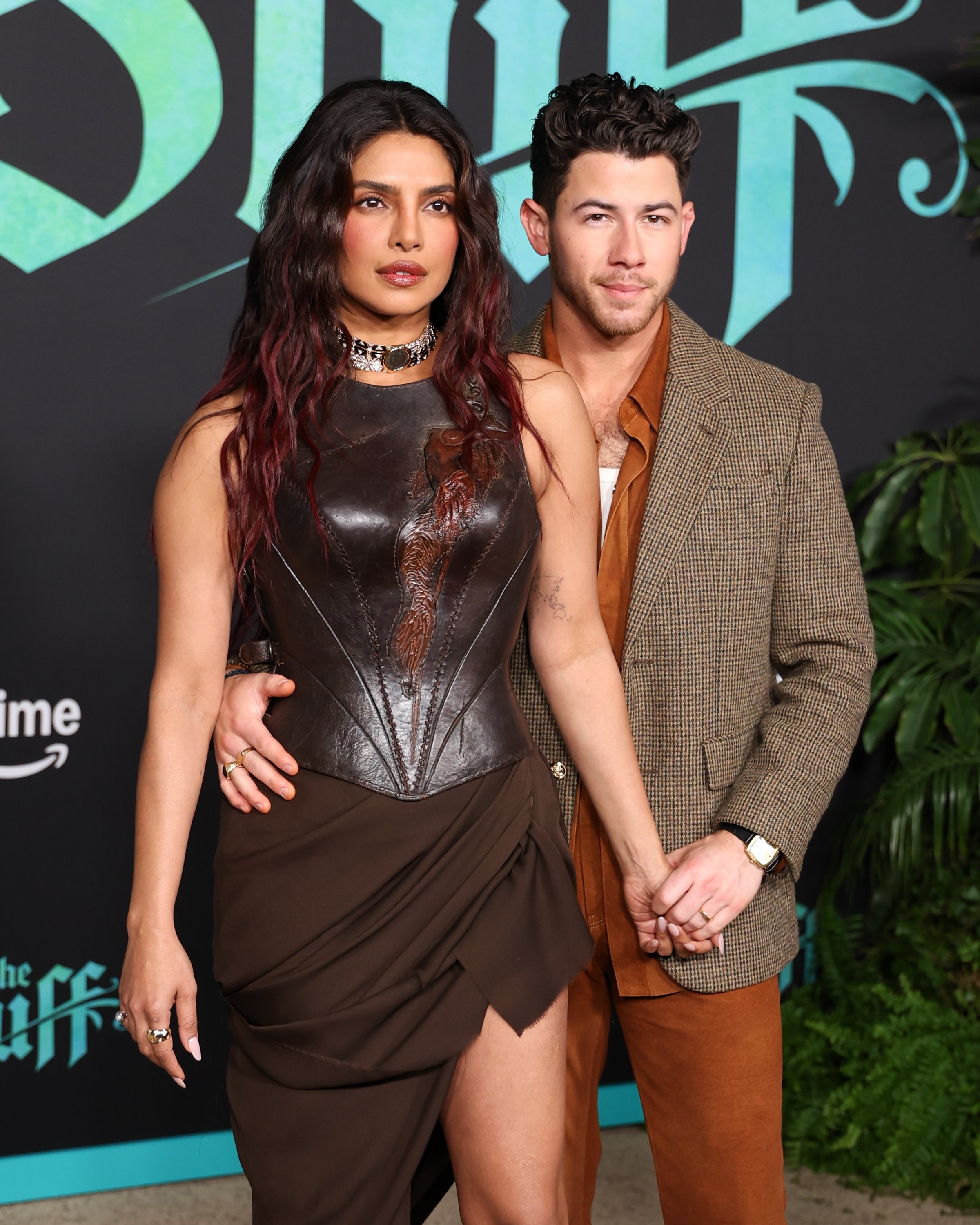 A woman in a leather corset top and skirt stands with a man in a brown suit holding hands, posing at a celebrity event