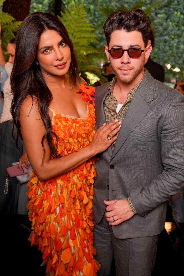 Two celebrities pose together; one in a ruffled, textured dress, the other in a plaid shirt and gray suit with sunglasses