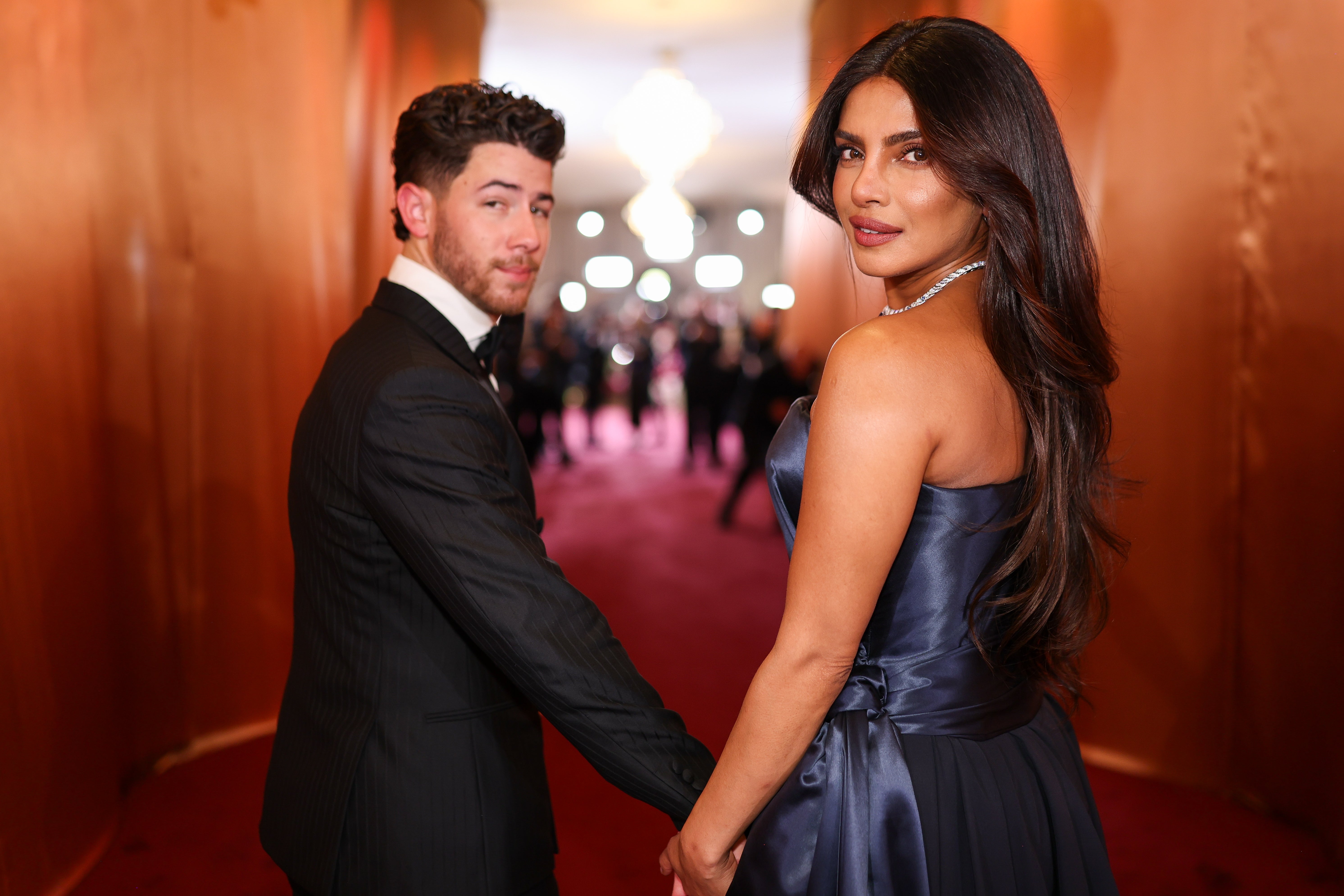 A couple on the red carpet holding hands; man in a suit, woman in an elegant gown with long hair and a necklace