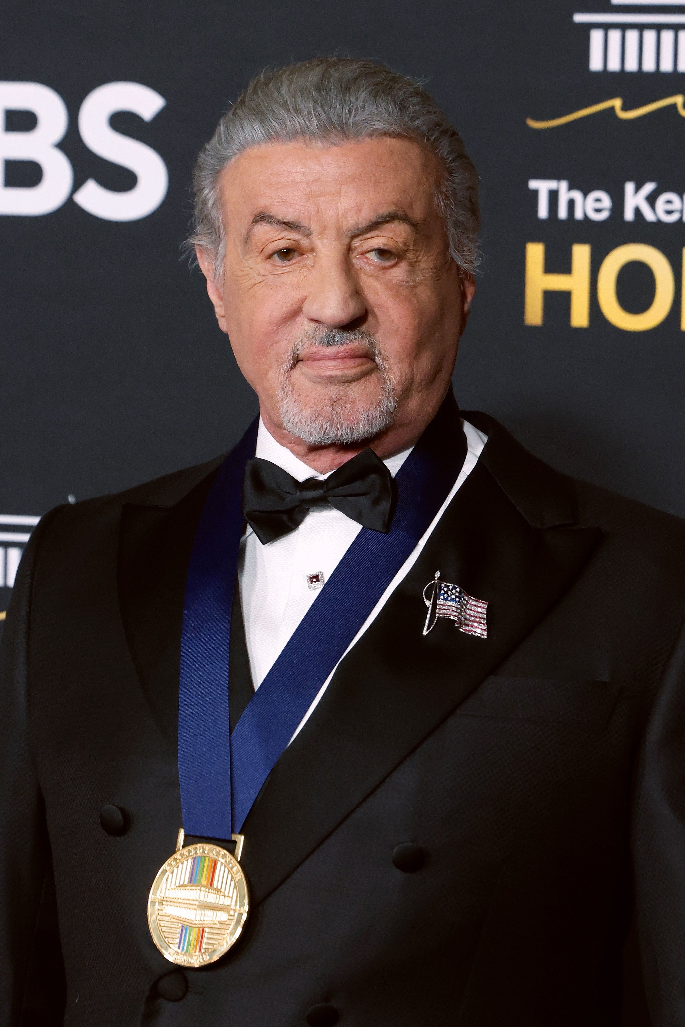 A man in a tuxedo with an American flag pin and a medal stands at an event