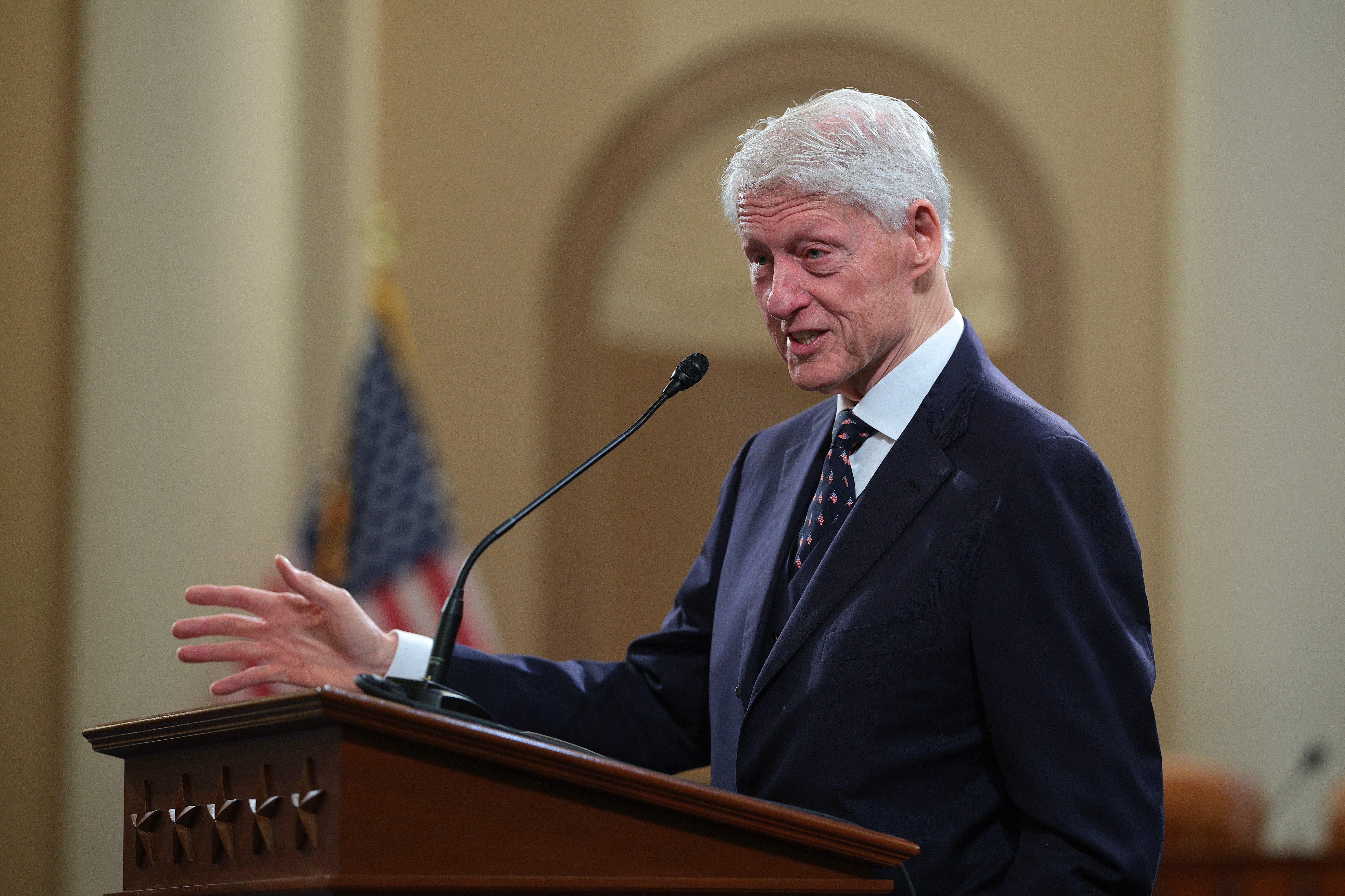 An unidentified person in a suit speaks at a podium with a microphone, an American flag in the background